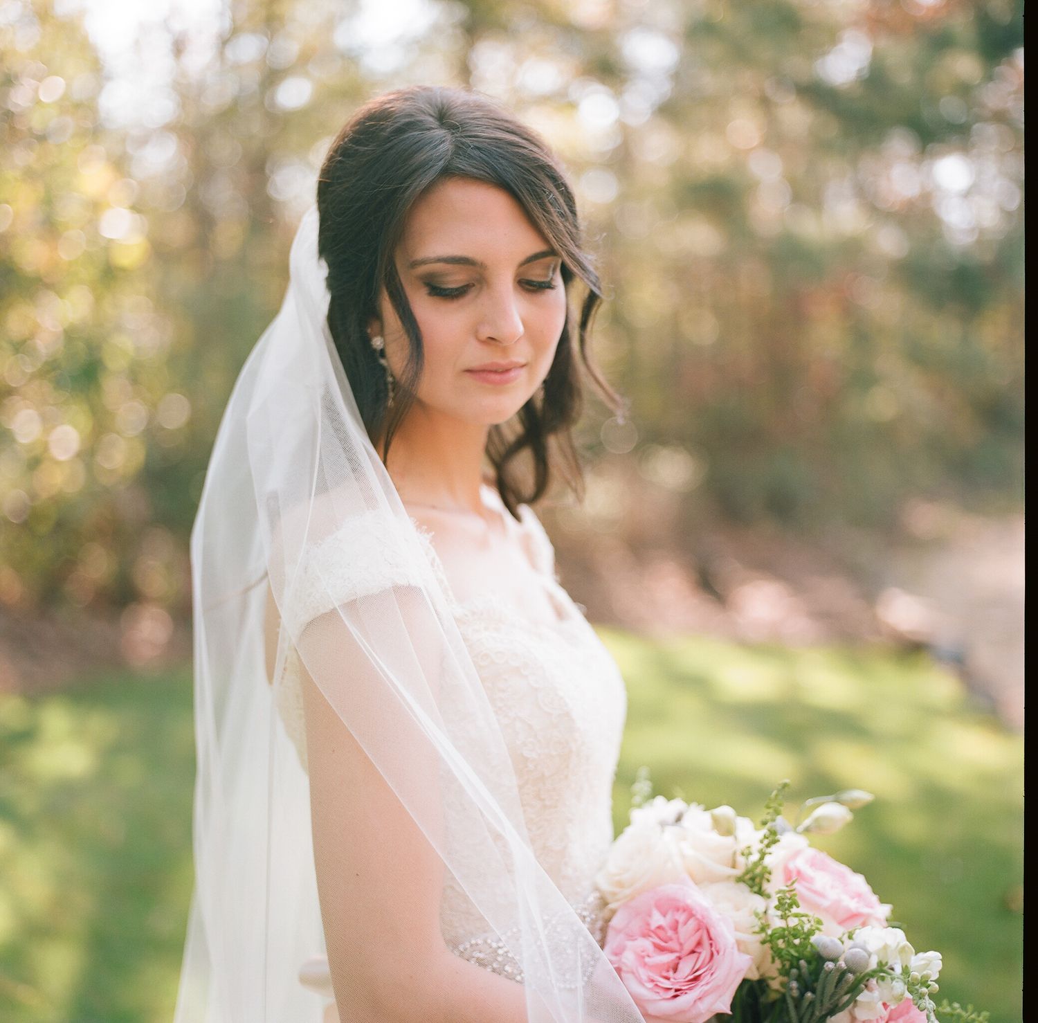 Bride holds pink and white bouquet while wearing lace gown and veil in garden setting.