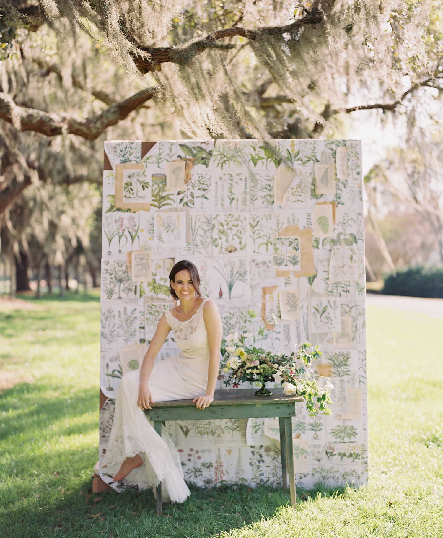 Bride in flowing white dress sits on green bench against stone wall with floral arrangements.