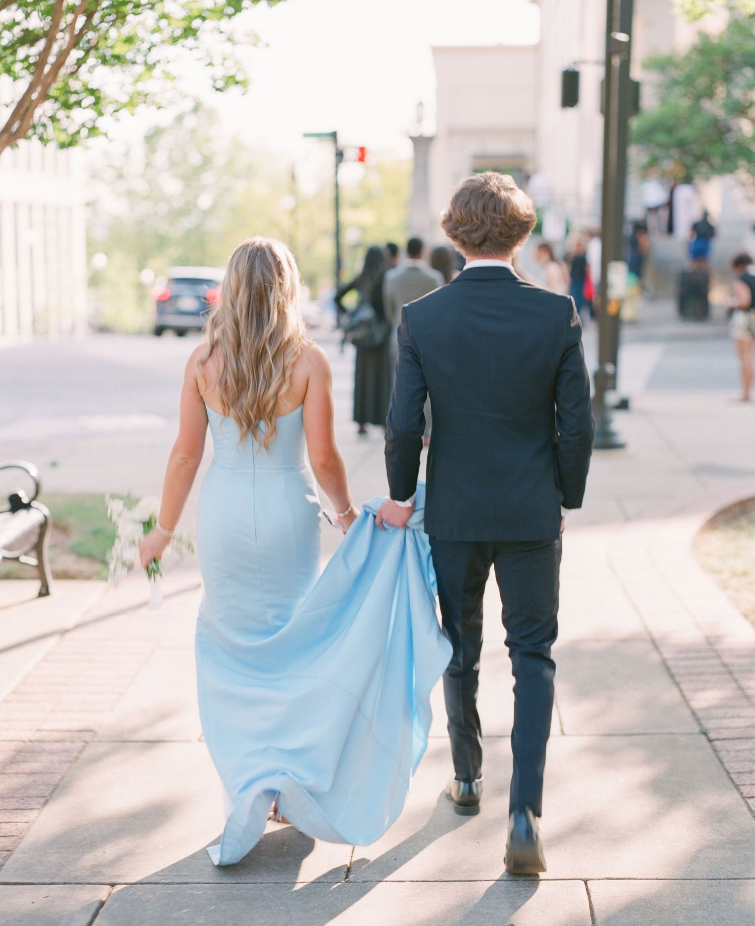 Couple walking hand-in-hand on sidewalk, man in navy suit carrying train of woman's light blue formal gown.