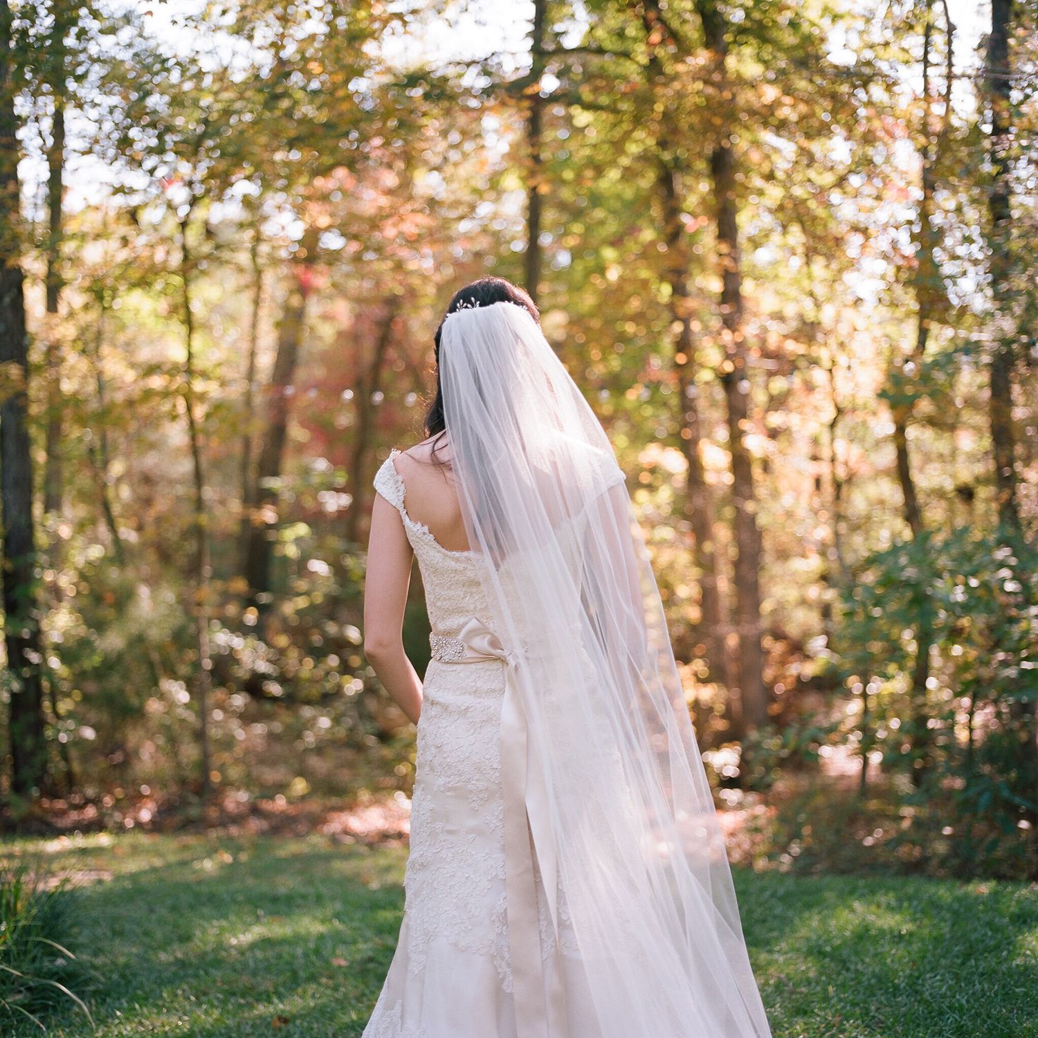 Bride in flowing white dress walks through autumn trees with trailing veil.