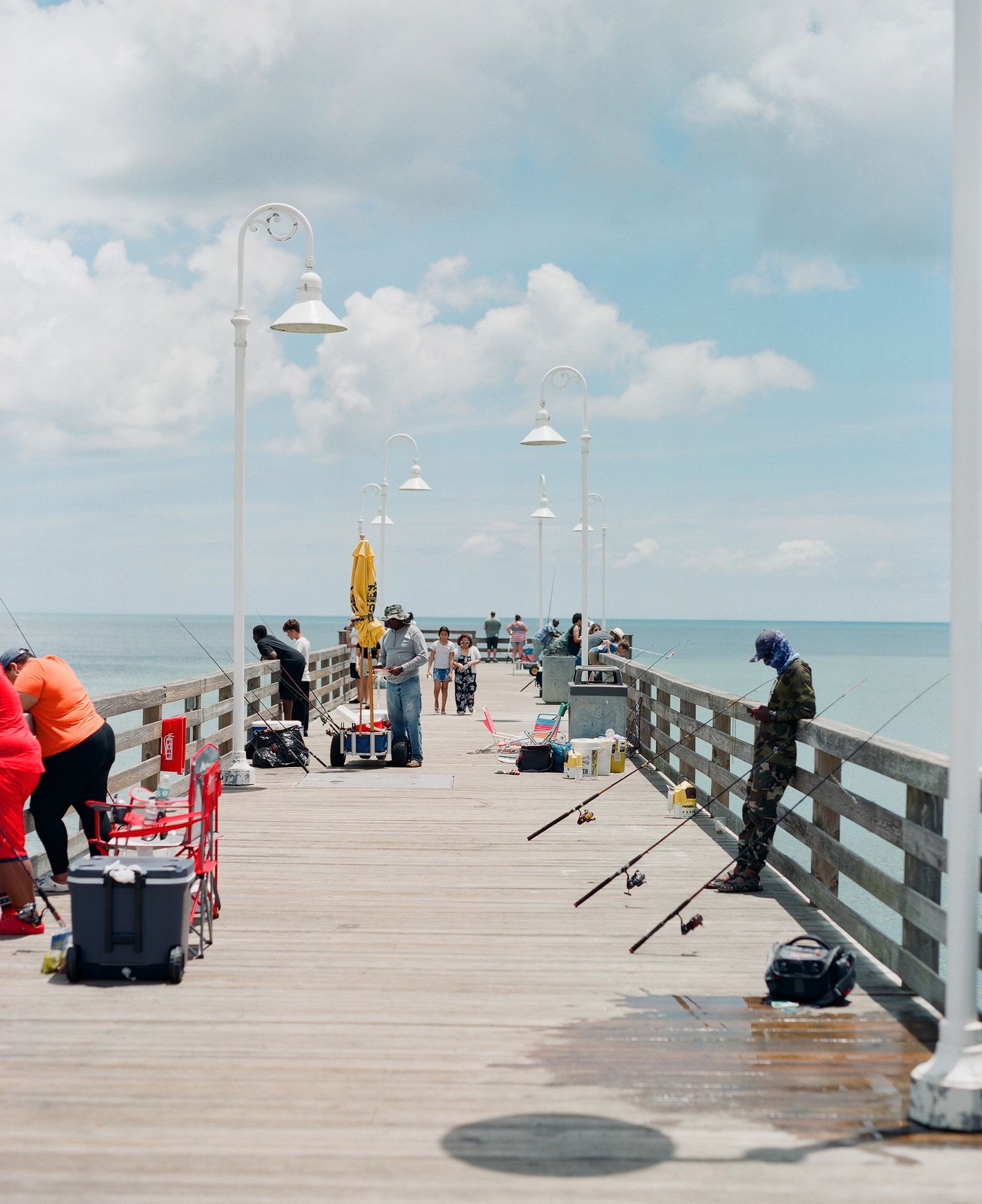 People fishing from a wooden pier on a sunny day with lamp posts and blue ocean views.