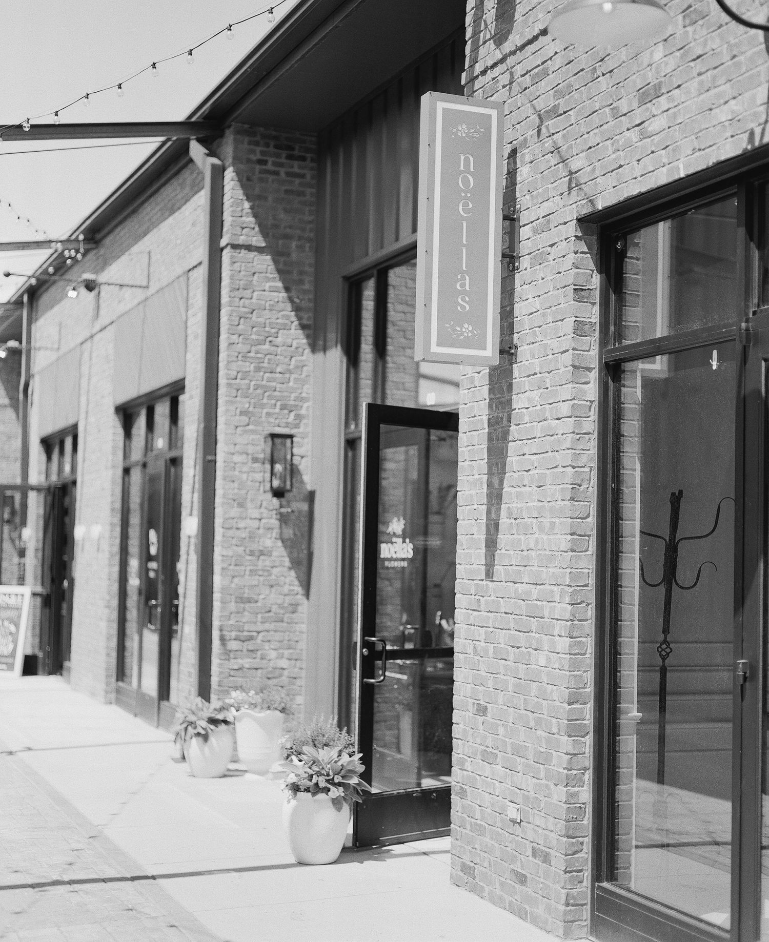 Black and white photo of a brick storefront with a hanging sign, potted plants, and string lights overhead.