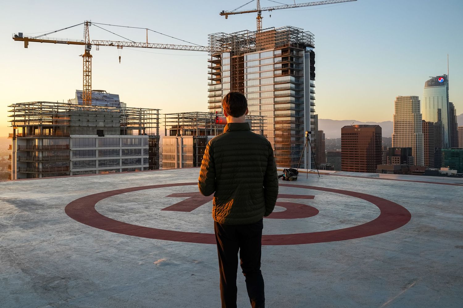 Silhouette overlooking construction cranes and city buildings from rooftop helipad at sunset.