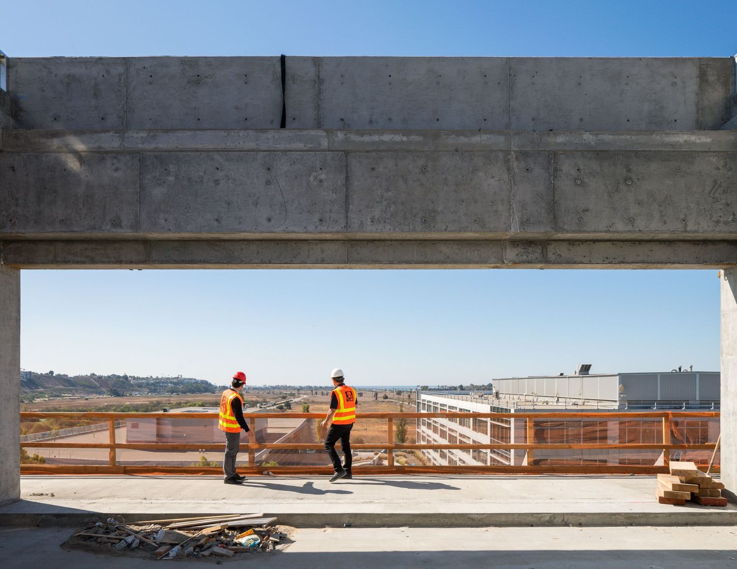 Construction workers in safety gear observe progress at a concrete building site overlooking an urban area.