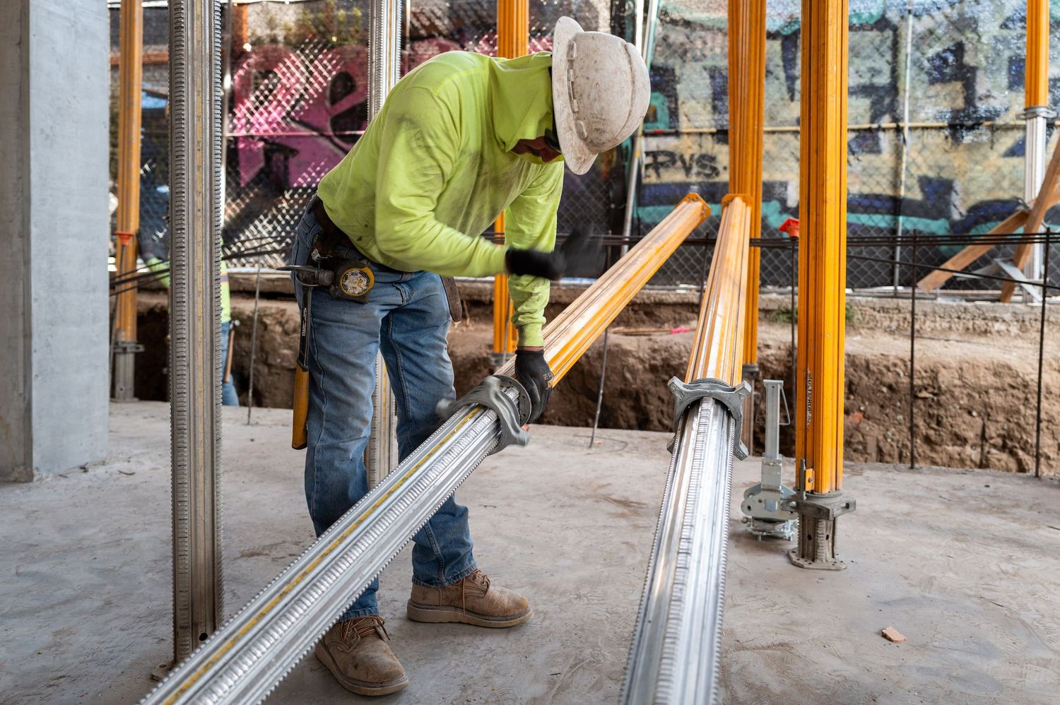 Construction workers installing yellow support beams and metal scaffolding in an unfinished building interior.