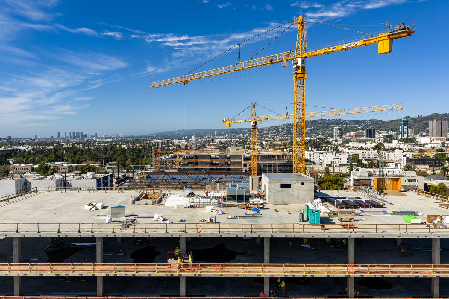 Construction cranes tower over a development site with blue sky and cityscape views in the background.