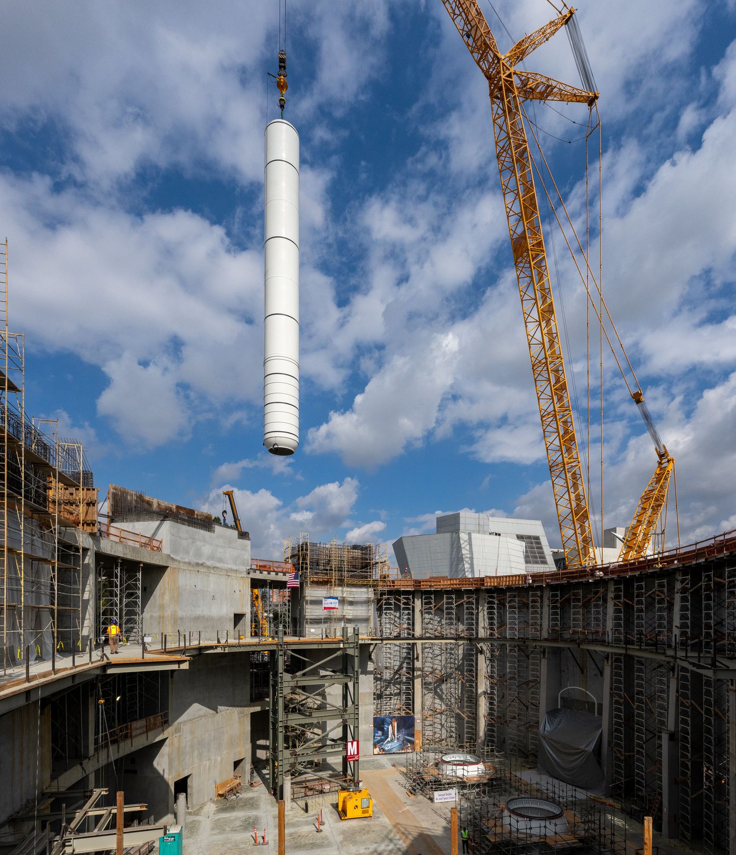 Large white cylindrical structure being lifted by crane at construction site with city backdrop.