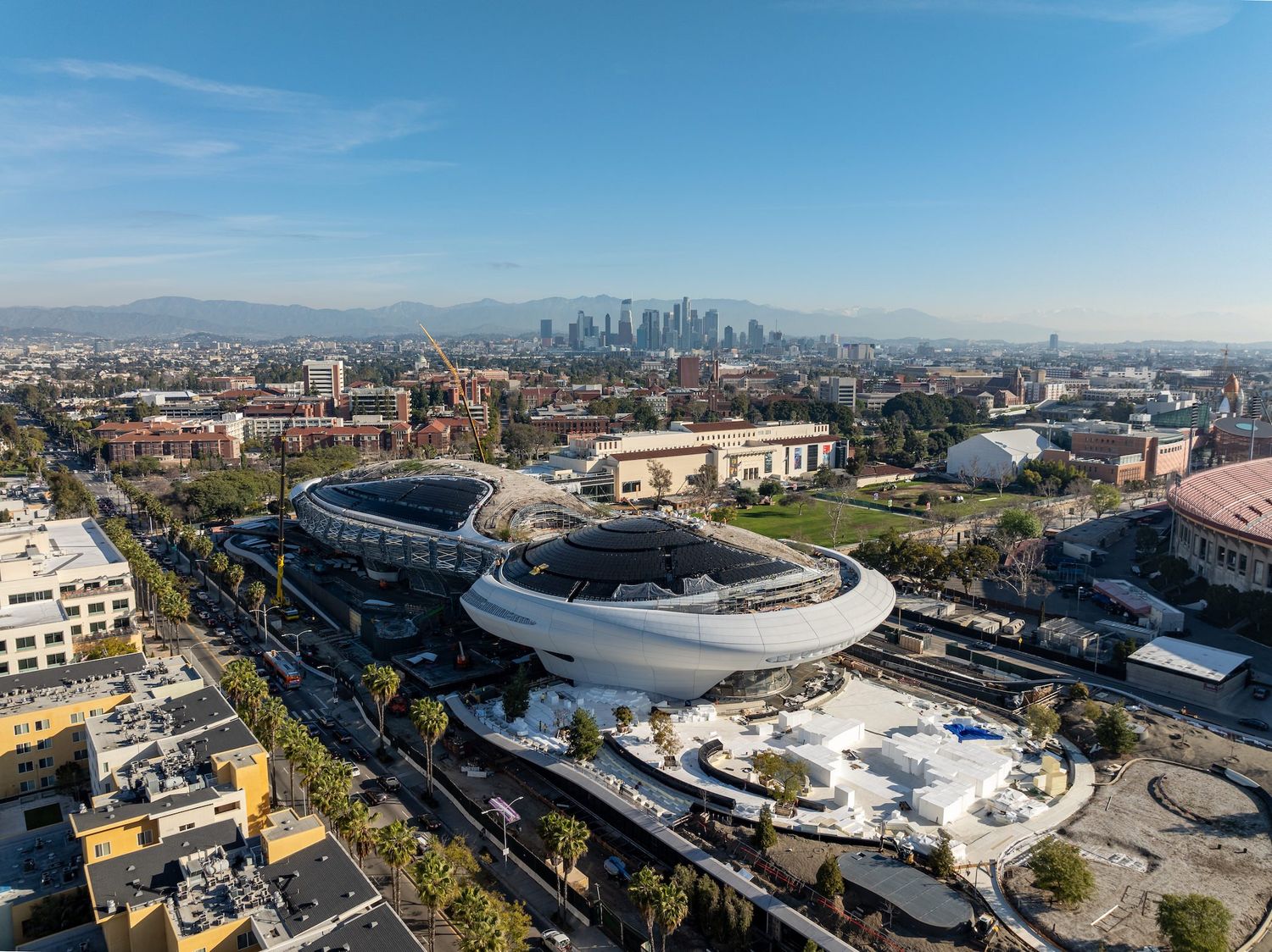 Sweeping aerial view of stadium construction site with Los Angeles downtown skyline visible in the distance.
