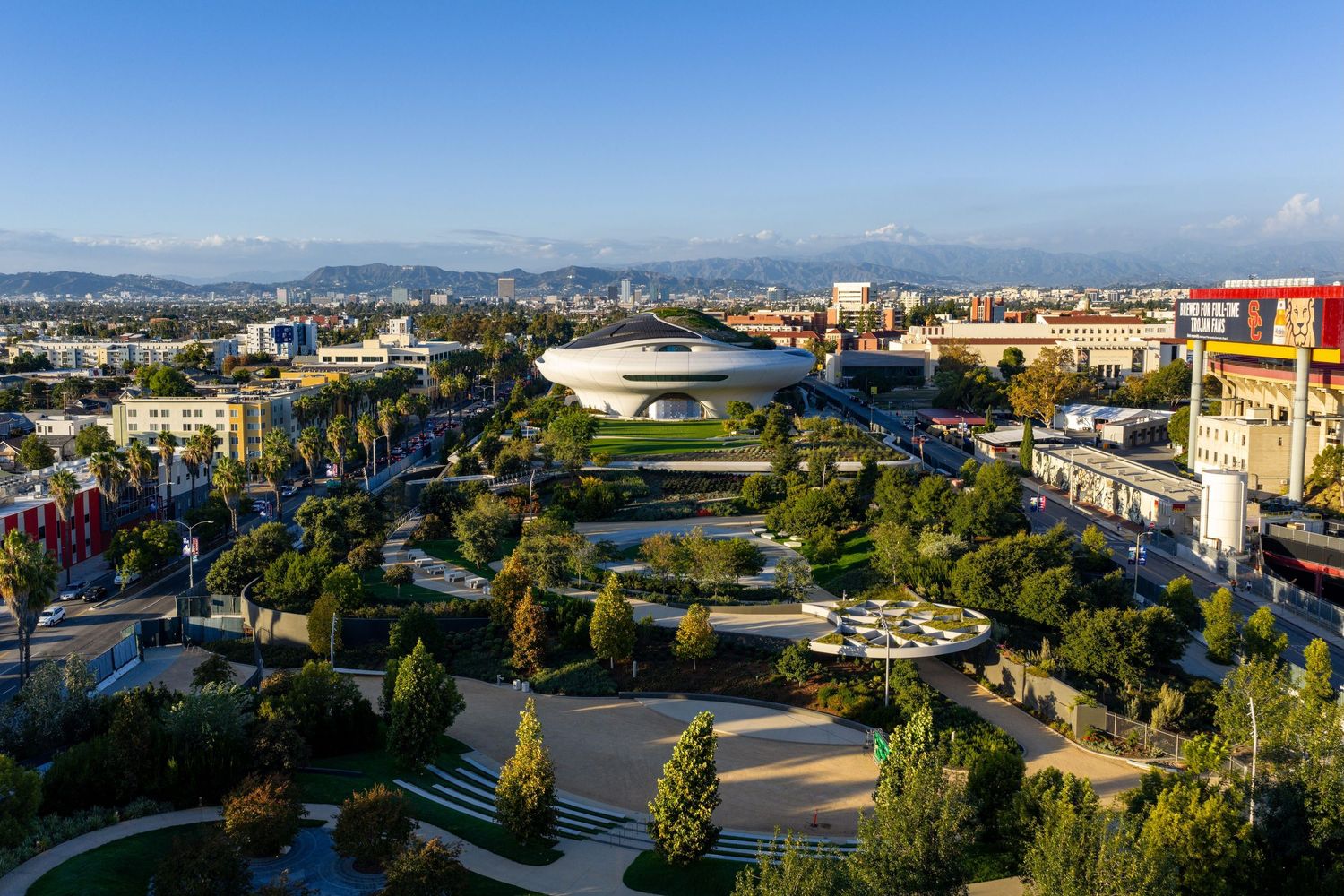 Aerial panoramic view of a modern stadium complex with curved architecture and surrounding cityscape.