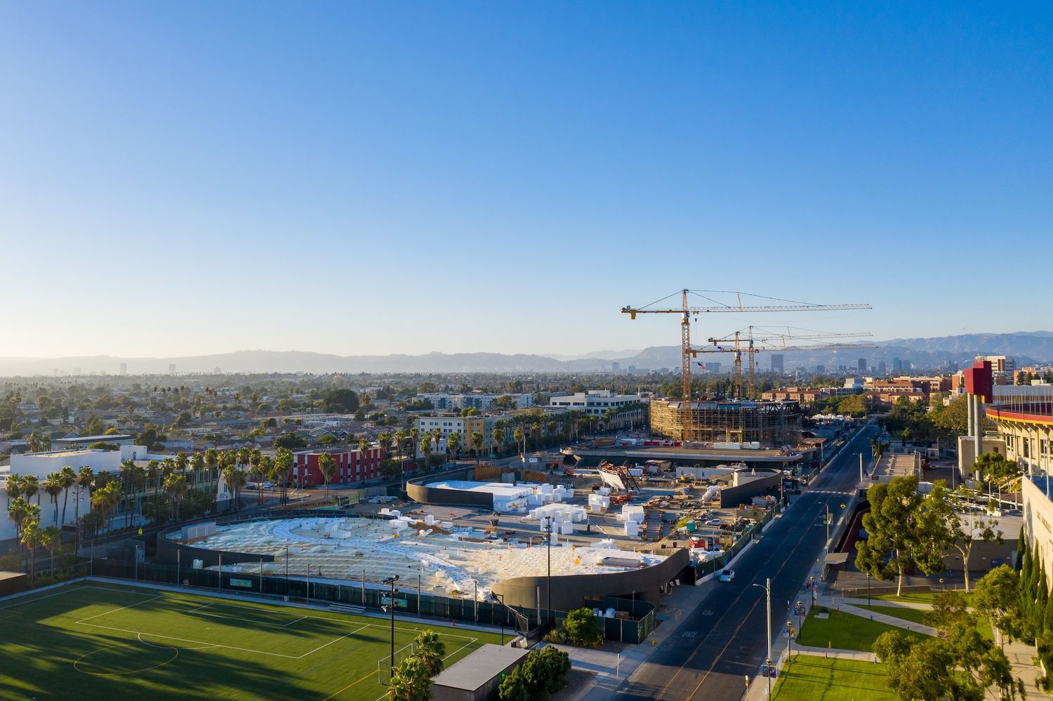 Wide aerial shot of a stadium development project with downtown Los Angeles skyline visible in the background.
