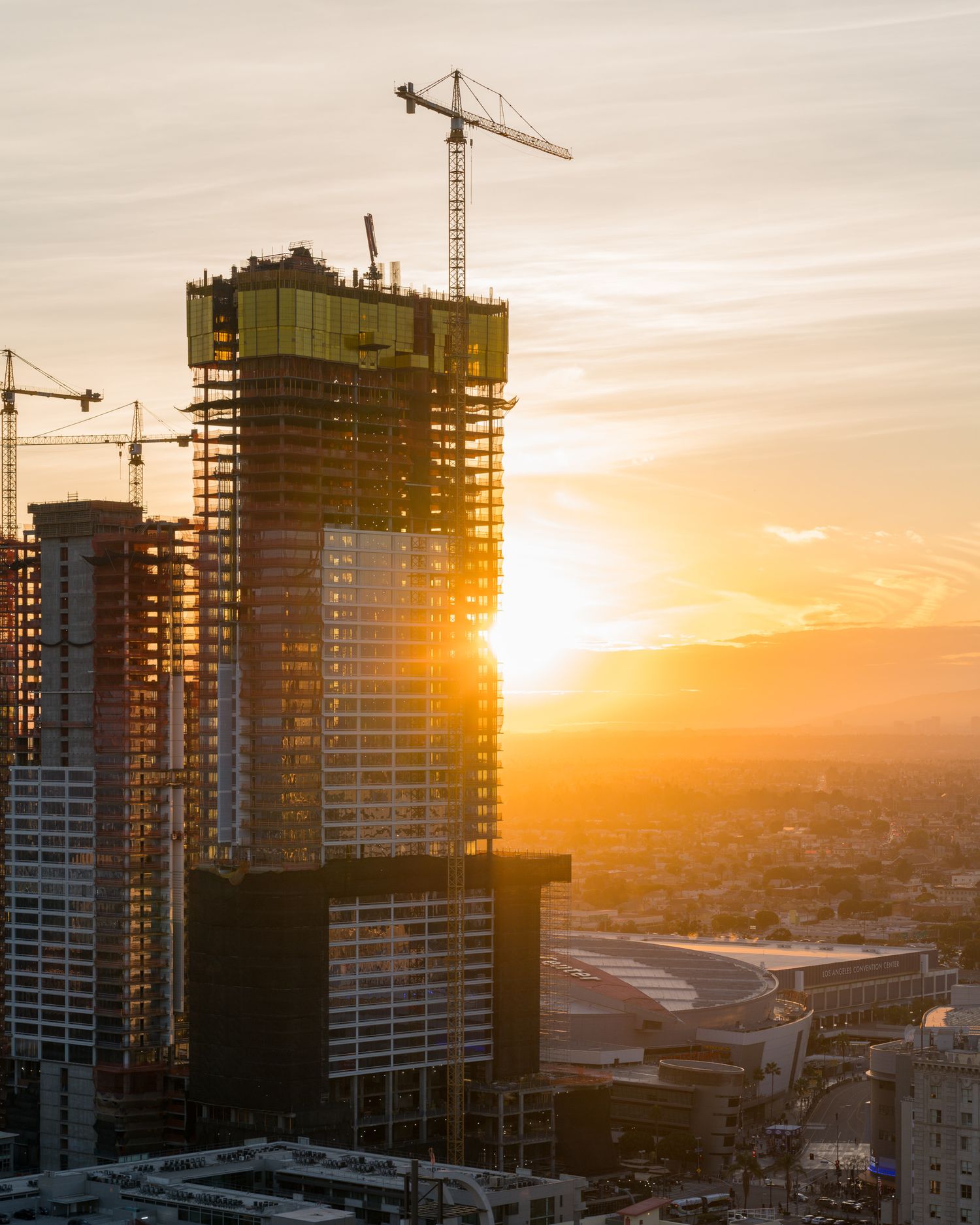 Construction cranes and high-rise buildings silhouetted against a dramatic orange sunset in an urban skyline.