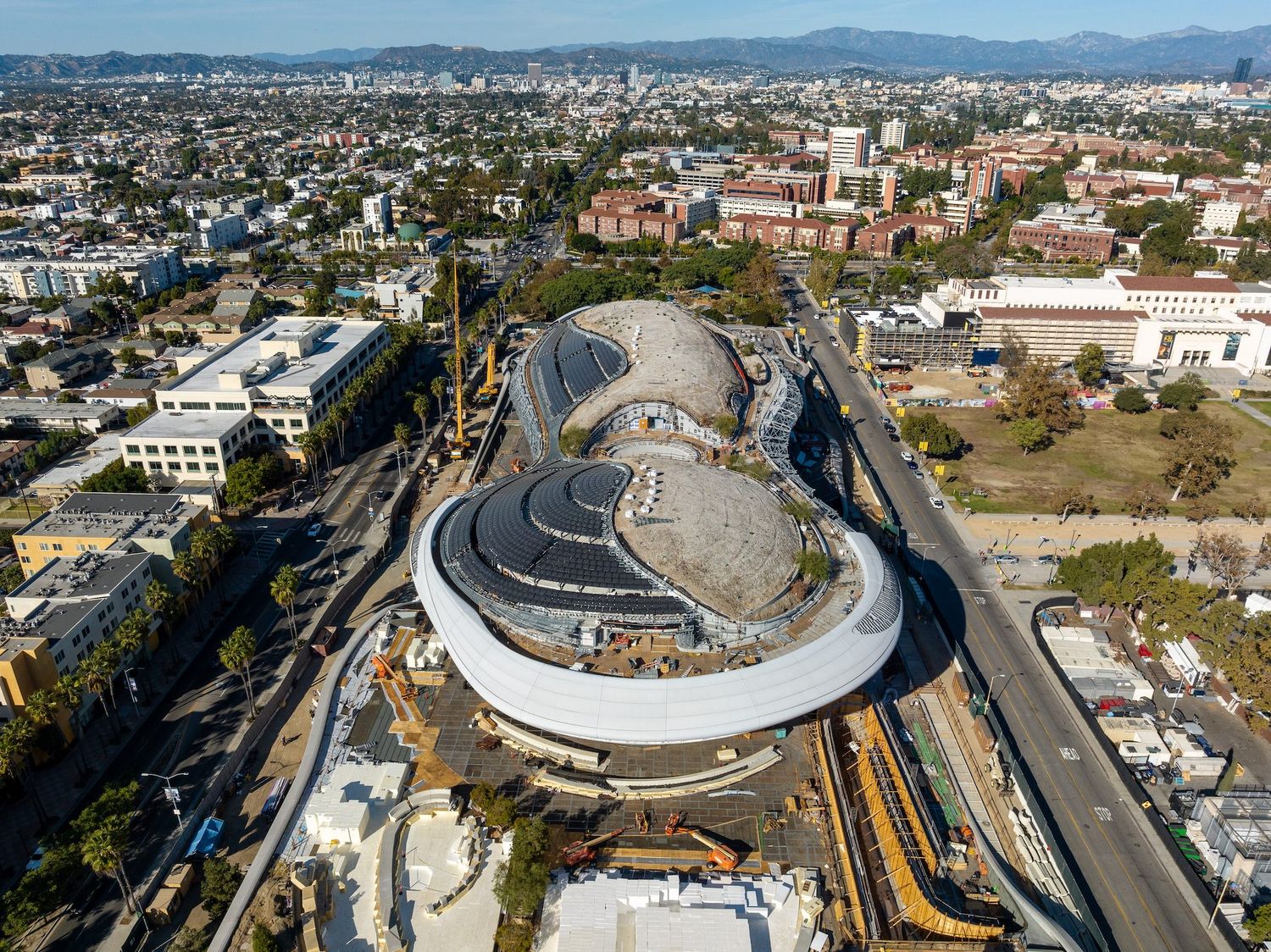 Aerial perspective of a modern stadium development with distinctive curved architecture and construction equipment.