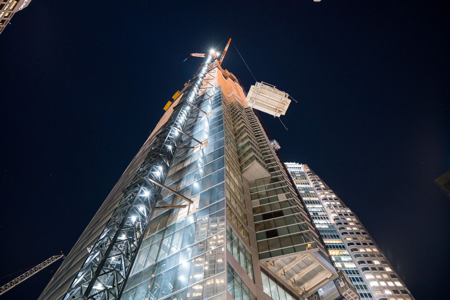Upward view of illuminated high-rise construction with cranes against night sky.