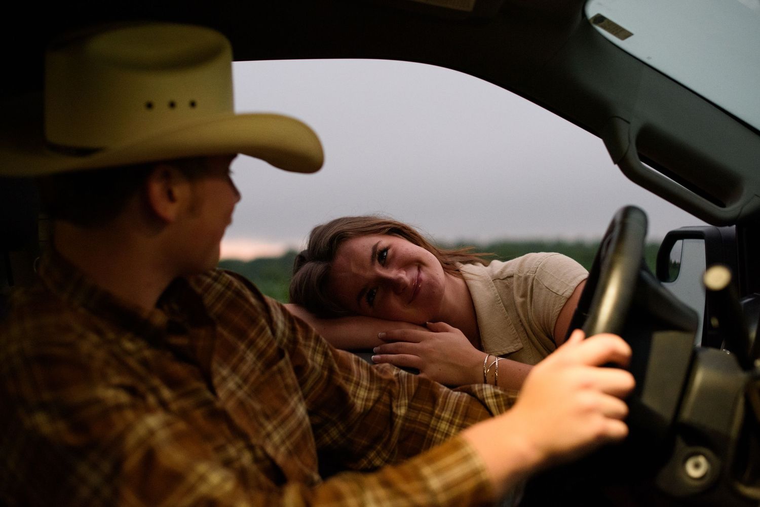 A woman rests her head on a man's shoulder in a truck cab, as he wears a cowboy hat and plaid shirt.