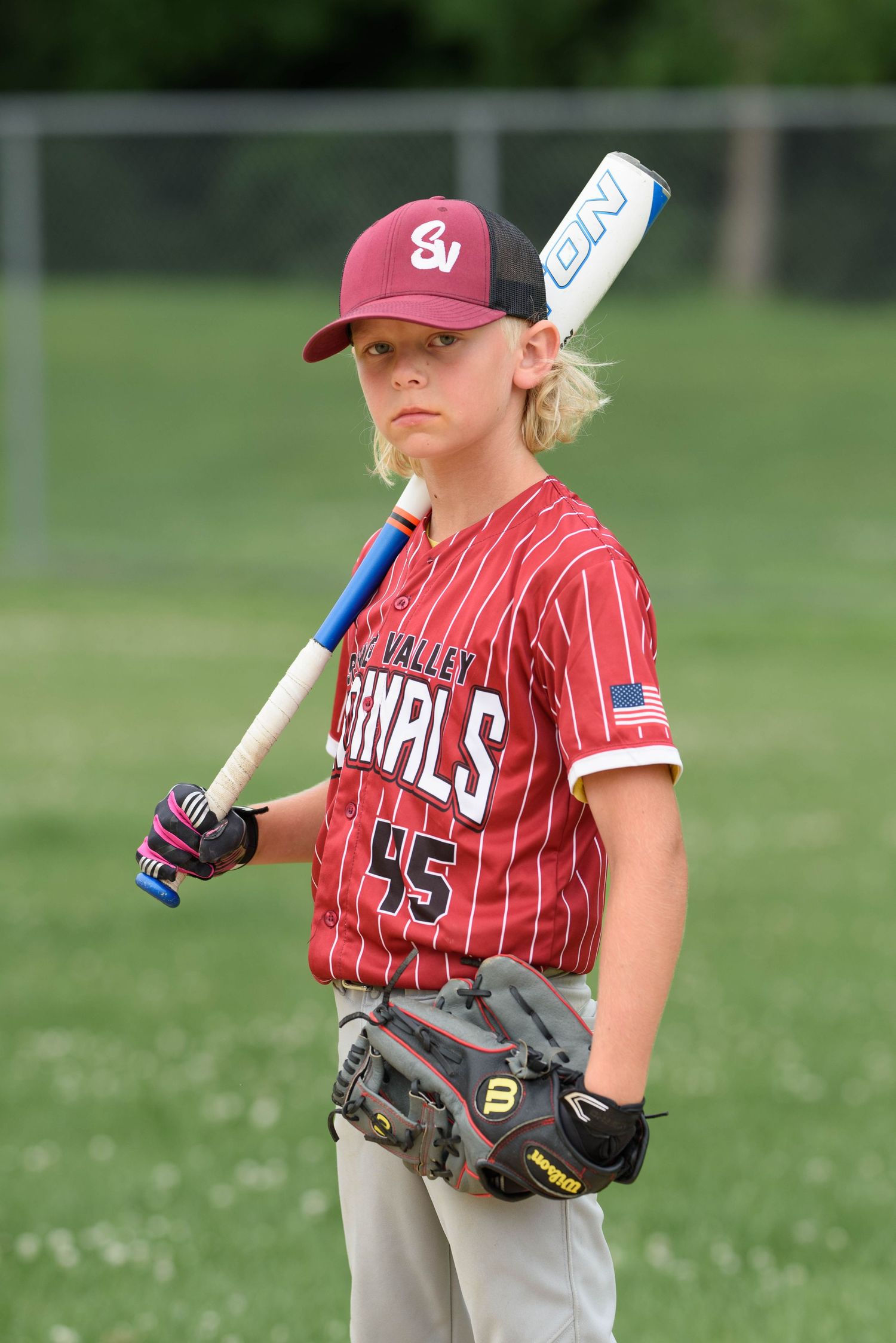 Young Cardinals baseball player #45 stands holding blue bat with glove in red pinstripe uniform outdoors.