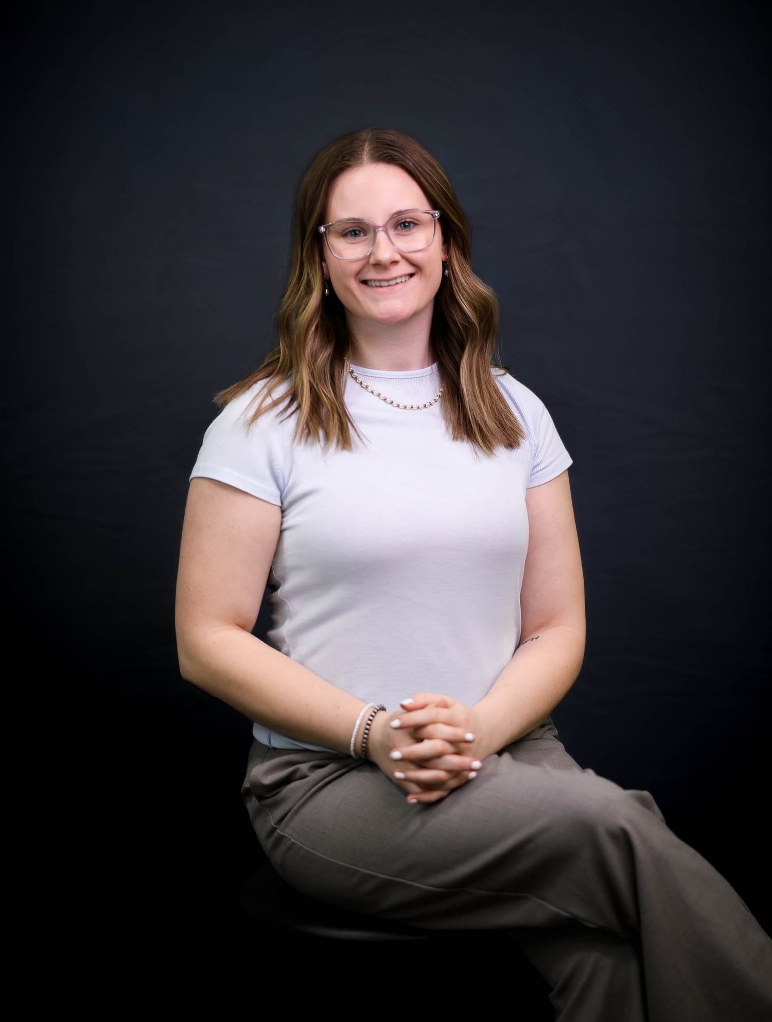 Young woman with glasses smiles in professional portrait wearing white top and gray pants.