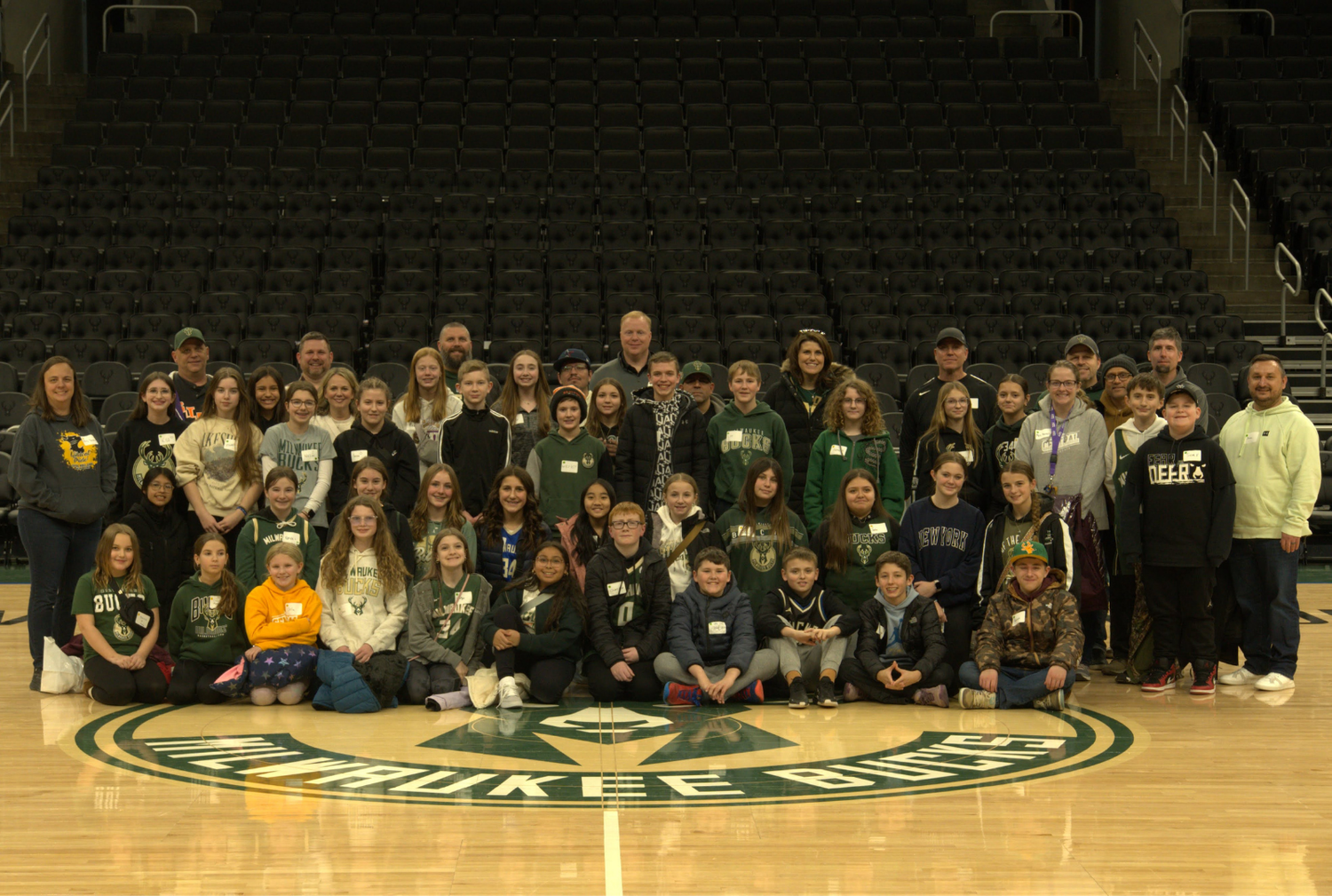 A large group of students and adults pose together on a basketball court logo at an arena.