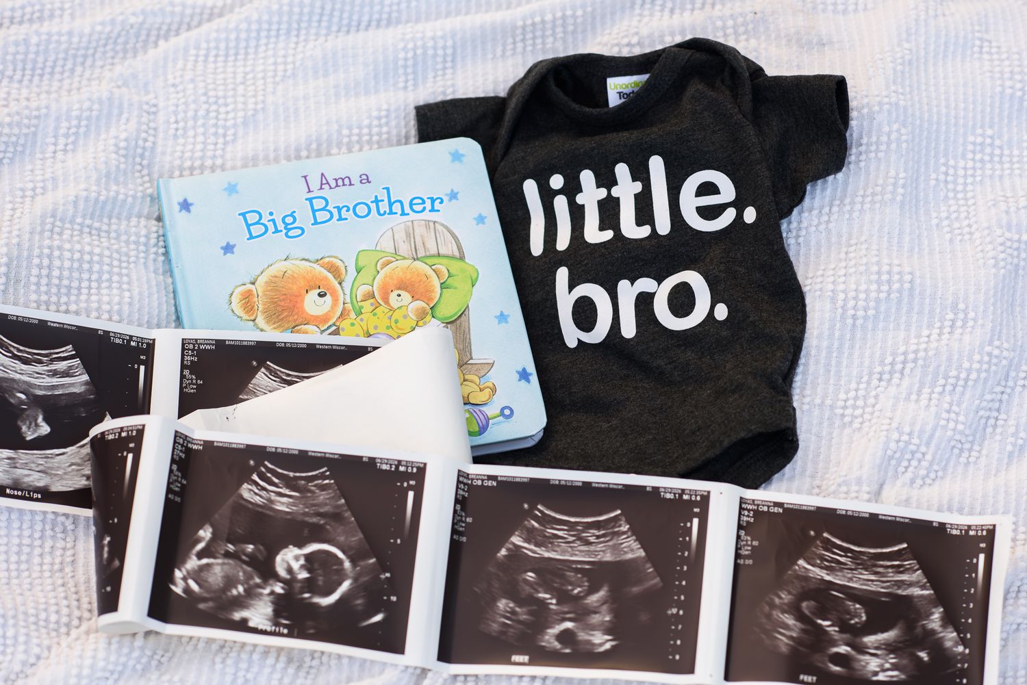 Baby gender reveal flat lay with ultrasound photos, little bro onesie, and I Am a Big Brother book on white background.