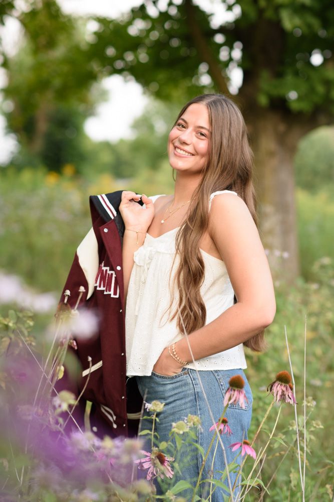 Smiling young woman with long hair holds a varsity jacket outdoors among wildflowers and green trees.