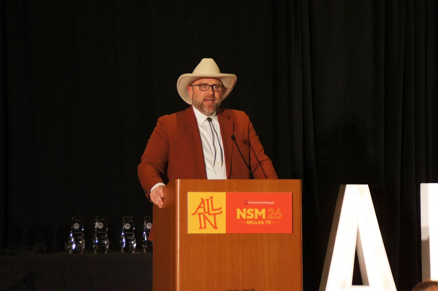 Man in cowboy hat and brown suit speaks at NSM 26 conference podium on stage.