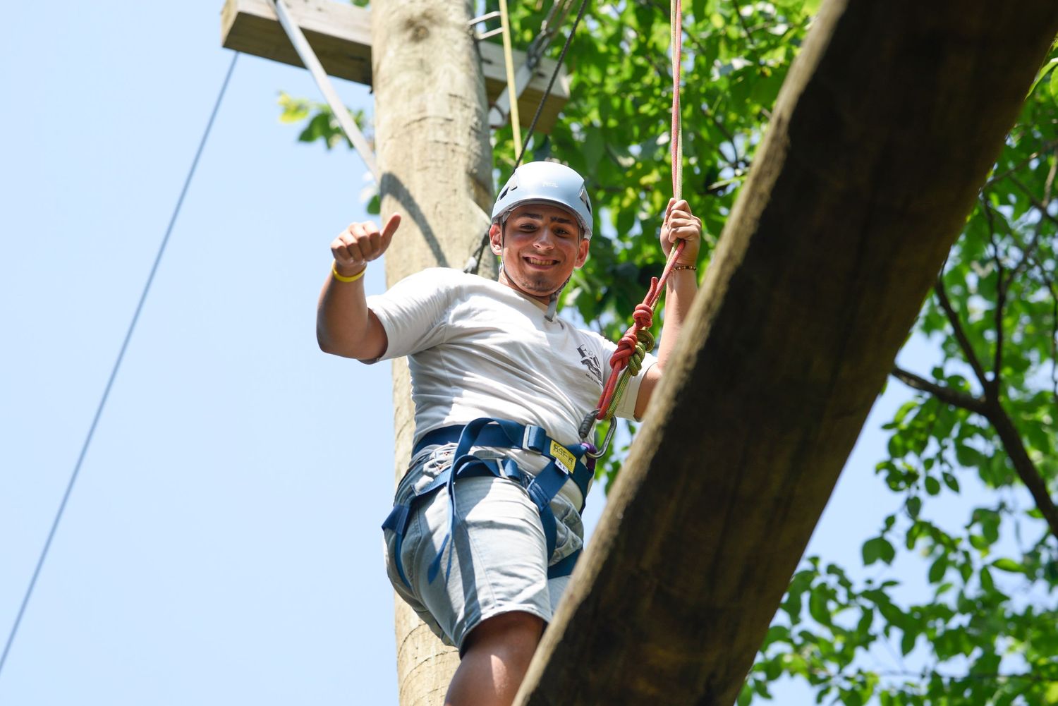 A smiling child wearing a helmet and harness climbs a wooden pole on a high ropes adventure course outdoors.