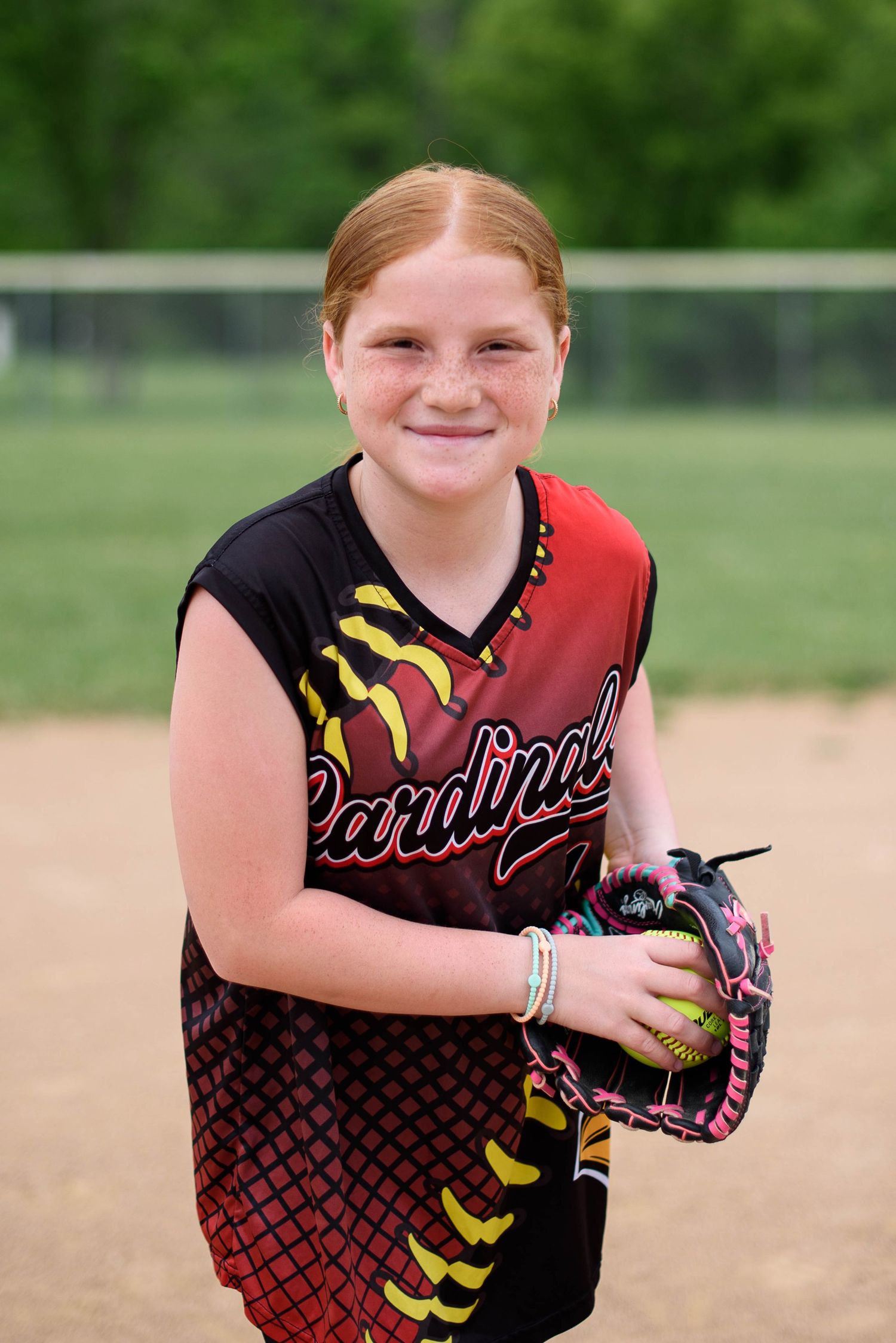 Young girl in Cardinals softball uniform holding glove and ball on dirt field.
