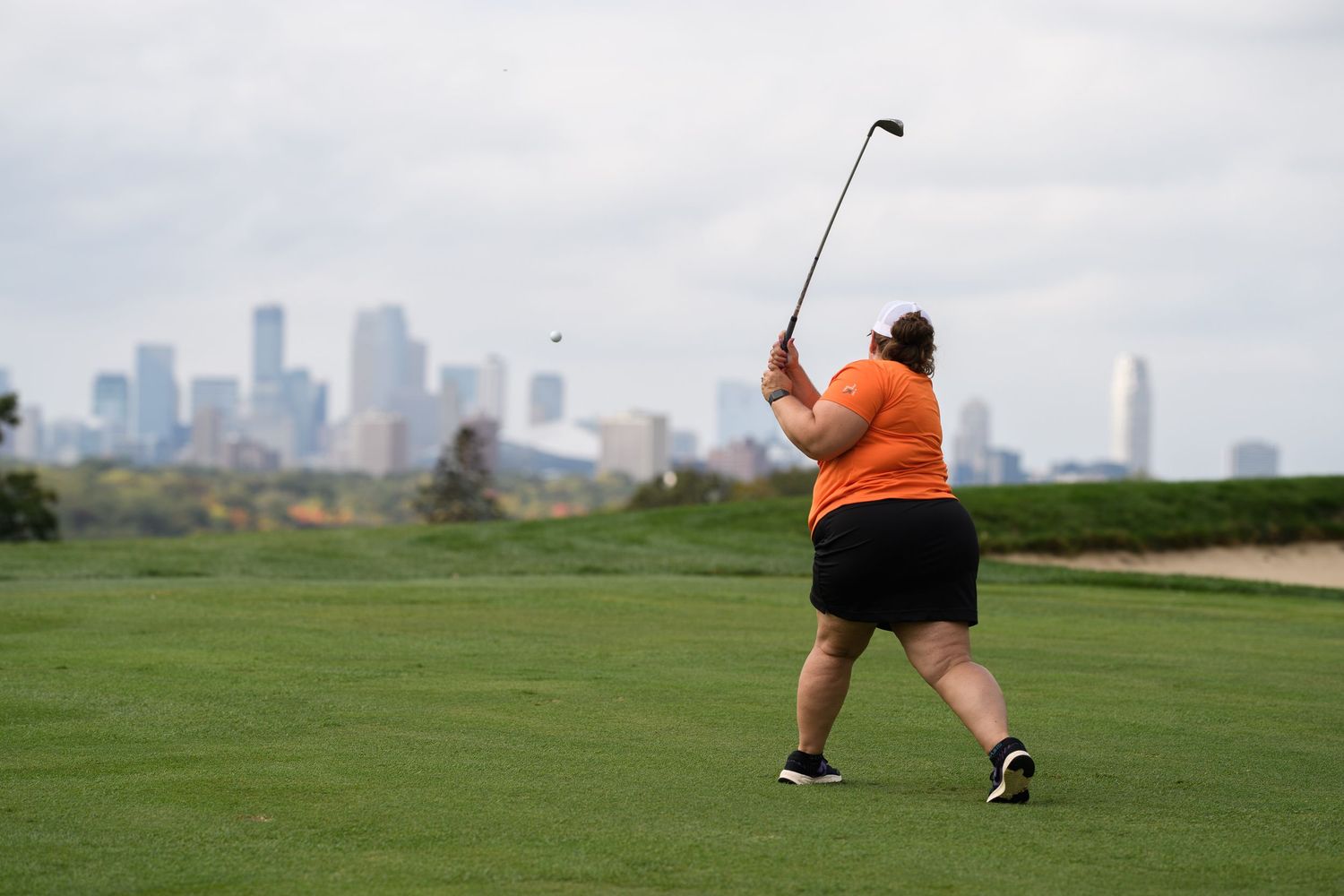 A golfer swings a club on a fairway with a city skyline visible in the background.