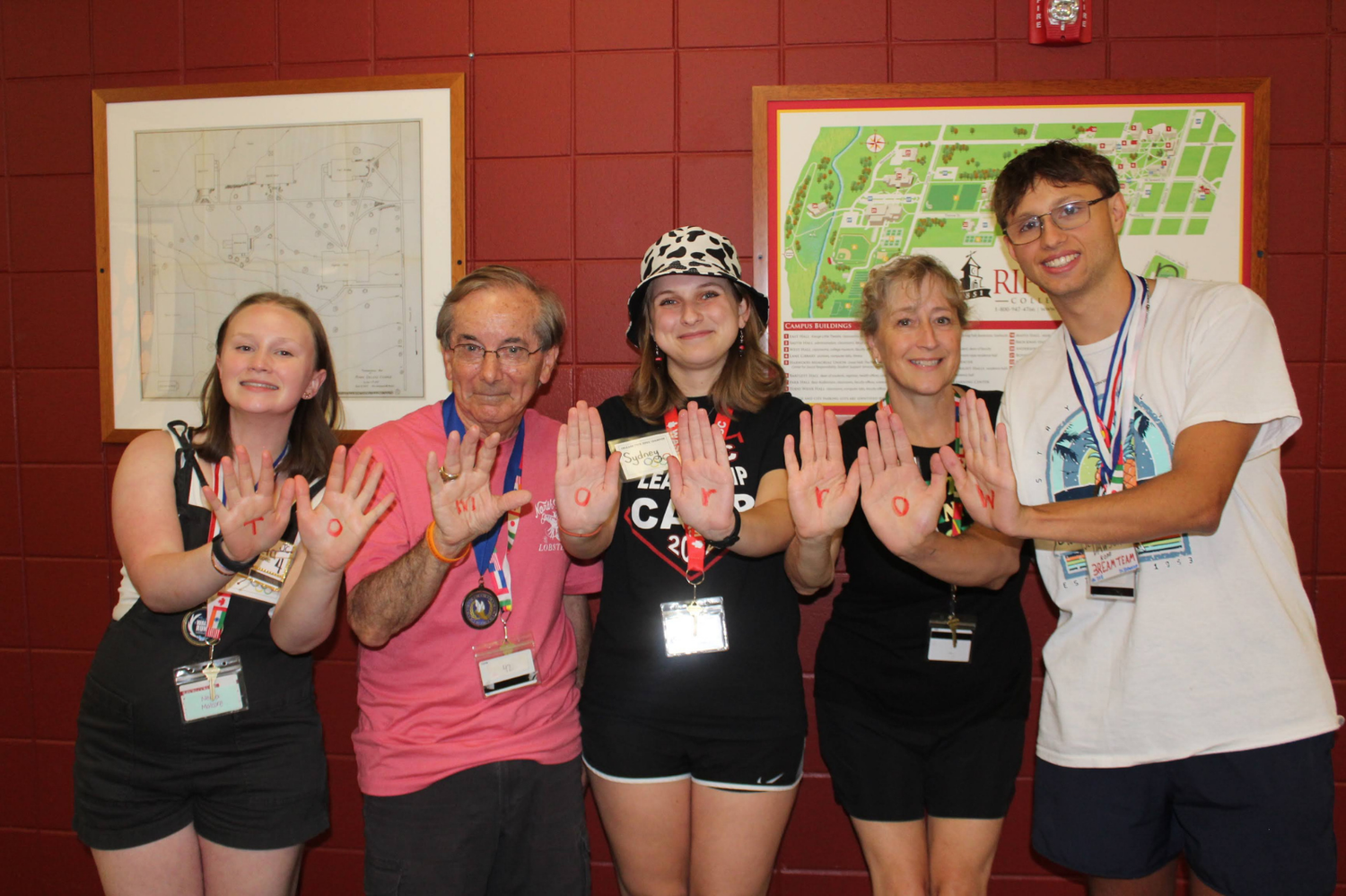 Five smiling camp participants pose with raised hands in front of a campus map on a red wall.