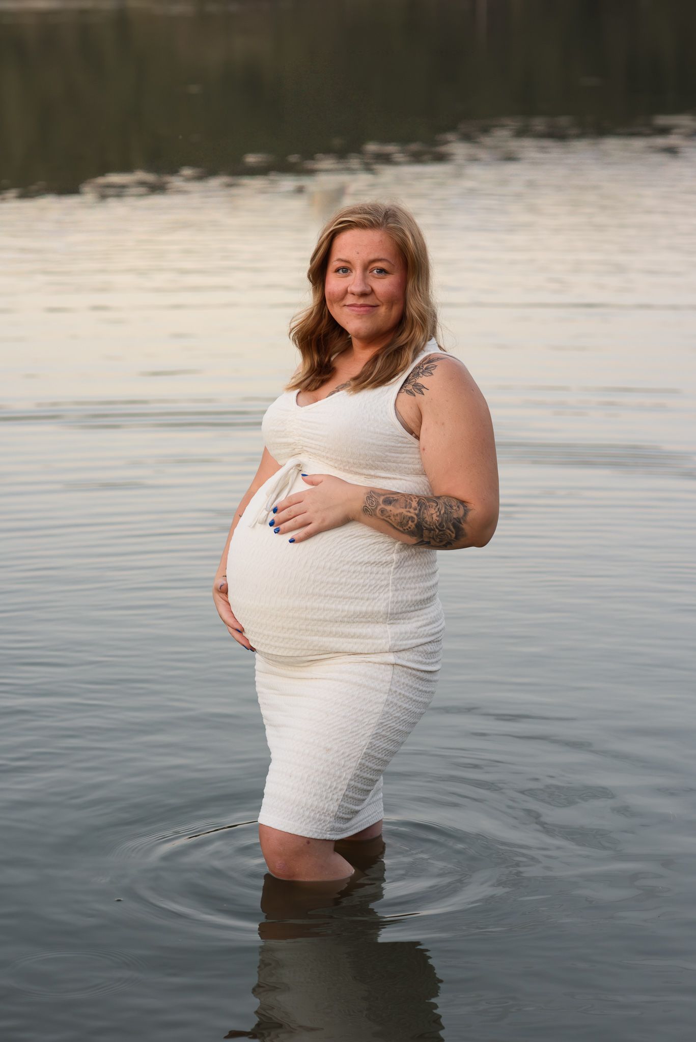 Pregnant woman in white dress standing in calm lake water, cradling baby bump, with tattoos visible on her arm.