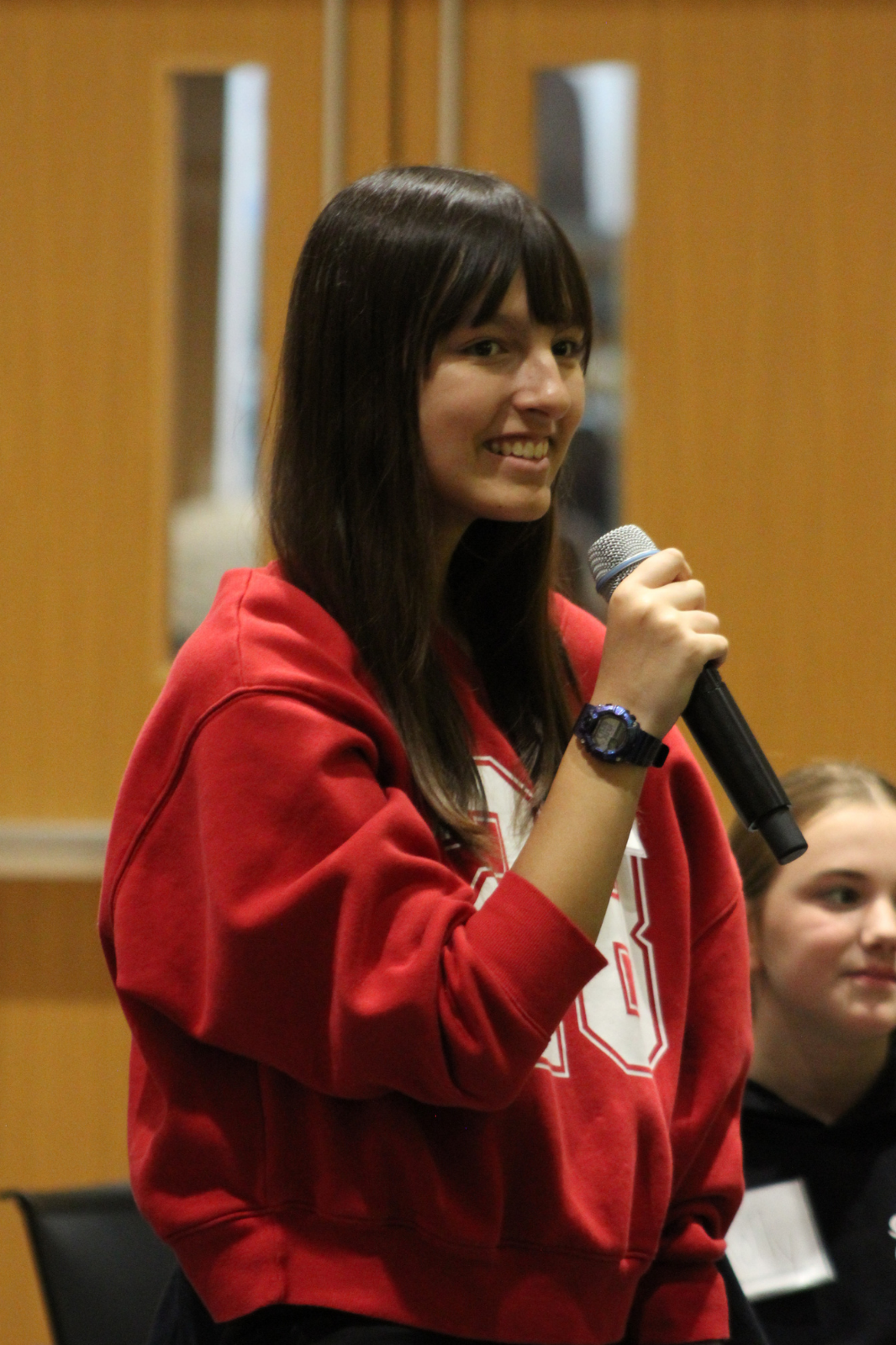 A smiling teenage girl in a red hoodie speaks into a microphone at an indoor student event.