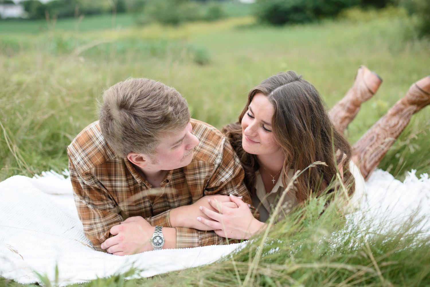 Couple lying on white blanket in green field, smiling at each other during romantic outdoor engagement photo session.