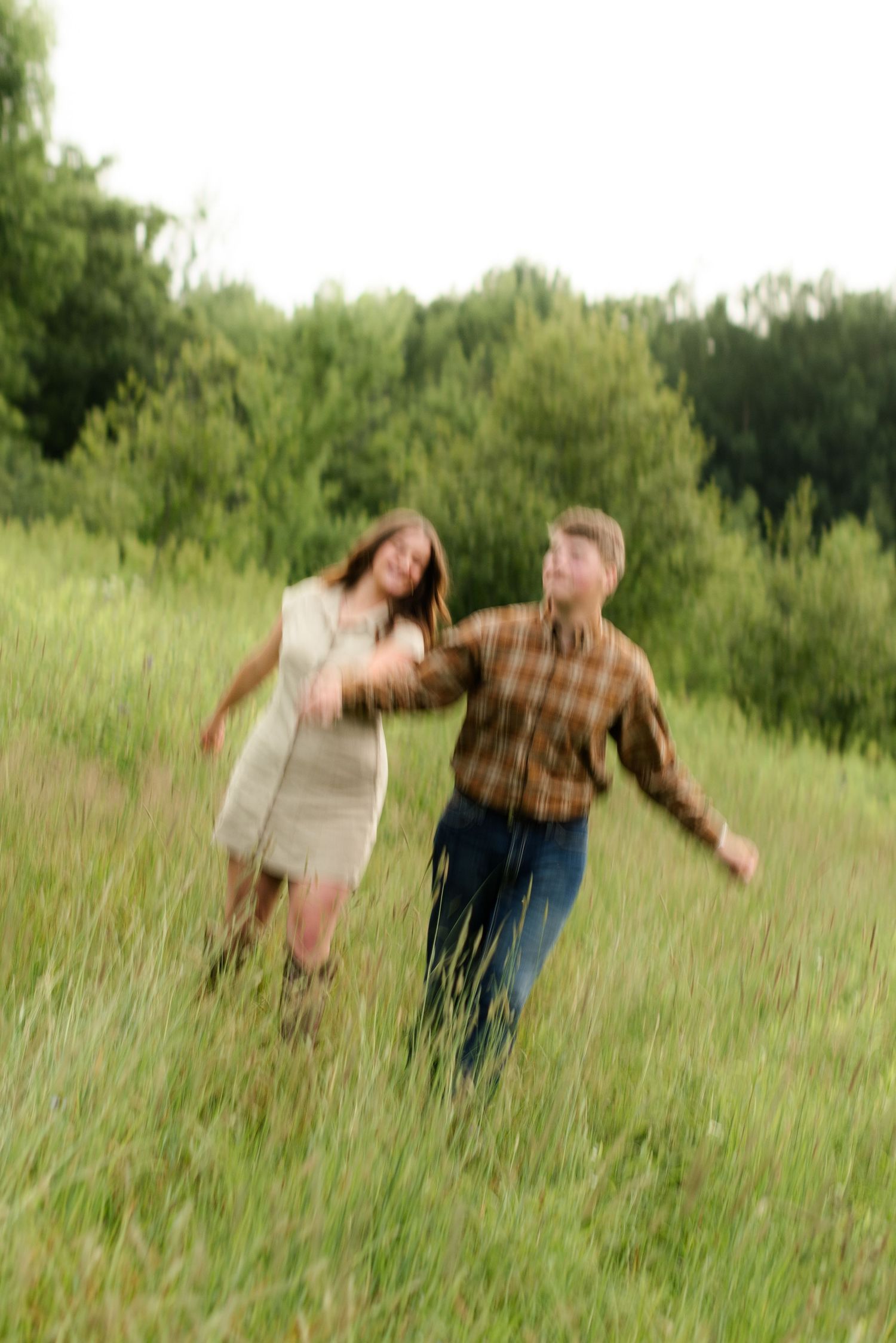 A couple joyfully runs through a lush green meadow, the woman in a cream dress and man in a plaid shirt.