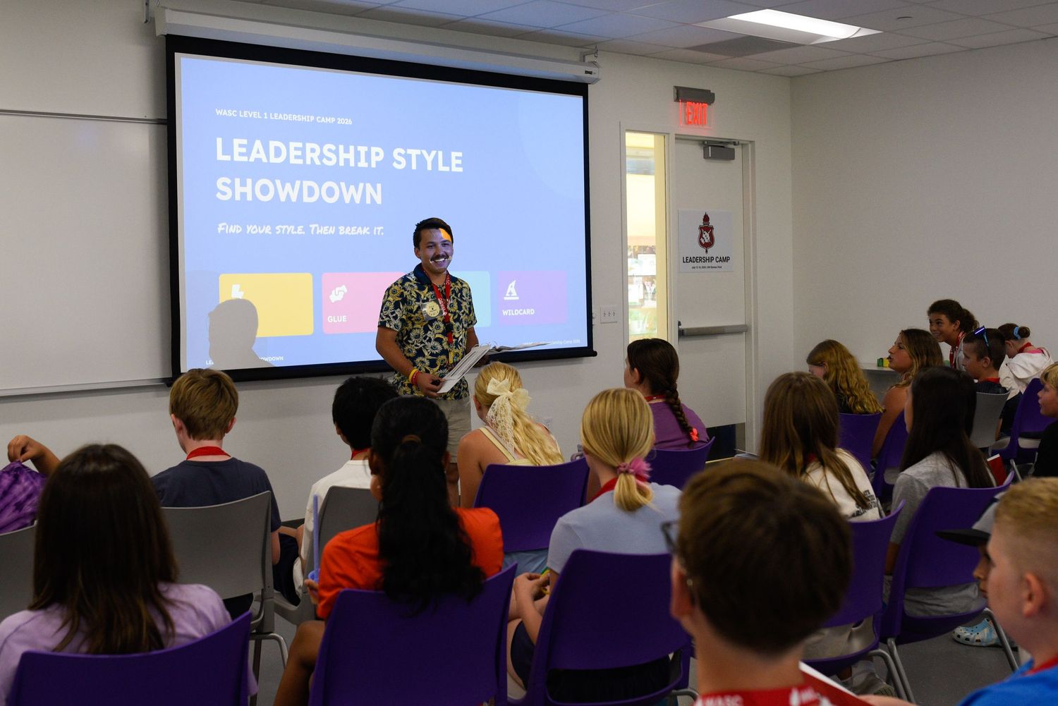 Speaker presenting Leadership Style Showdown presentation to students seated in purple chairs in a modern classroom.