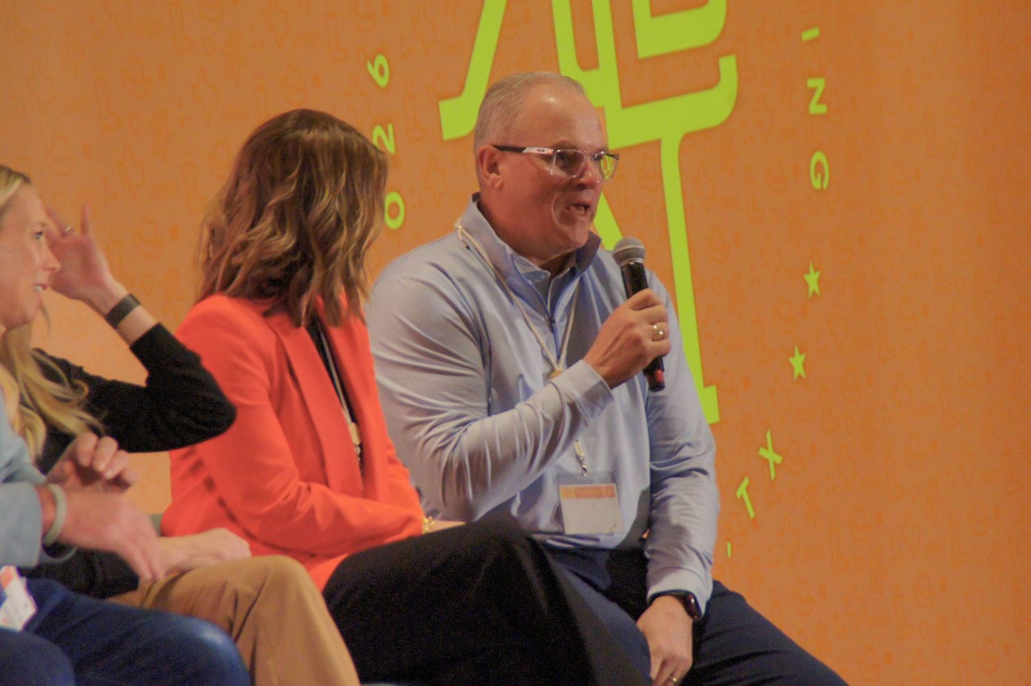 Man in light blue shirt speaks into microphone at a professional panel event.