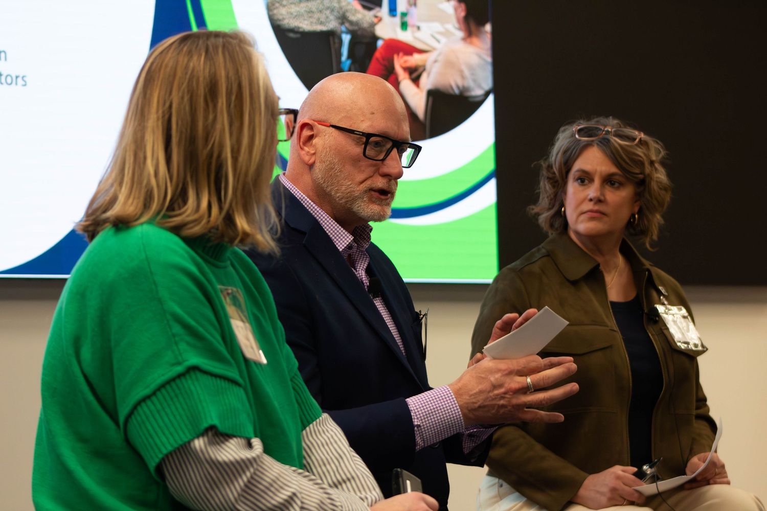Three professionals in discussion at a corporate event, with a green presentation screen visible in the background.