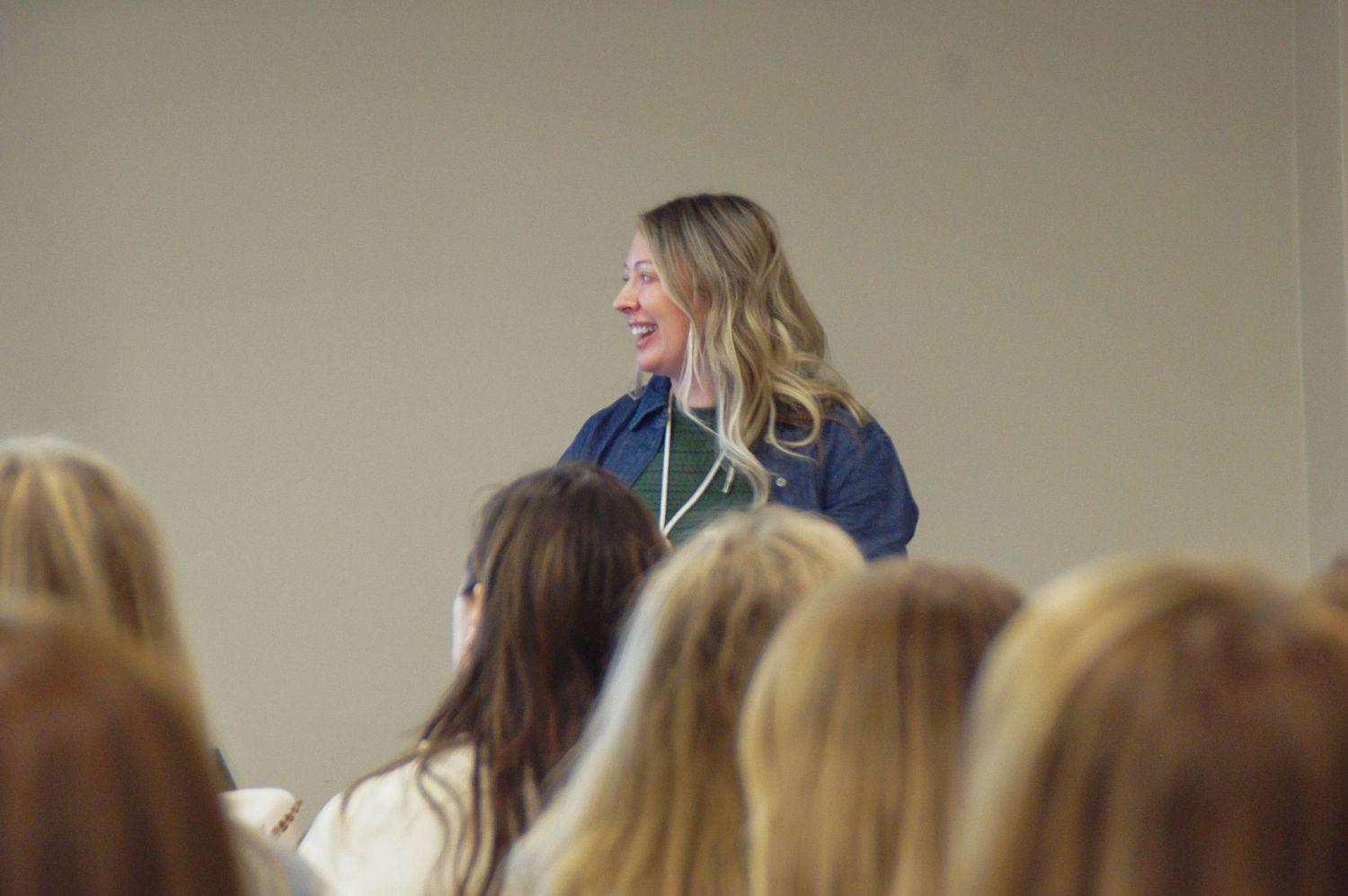 Blonde woman in blue blazer smiles while presenting to seated audience in conference room.