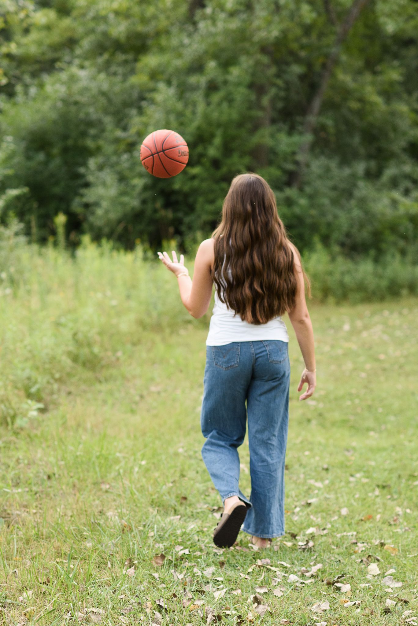 Young girl with long brown hair tossing a basketball in a green park, wearing white tank top and jeans.