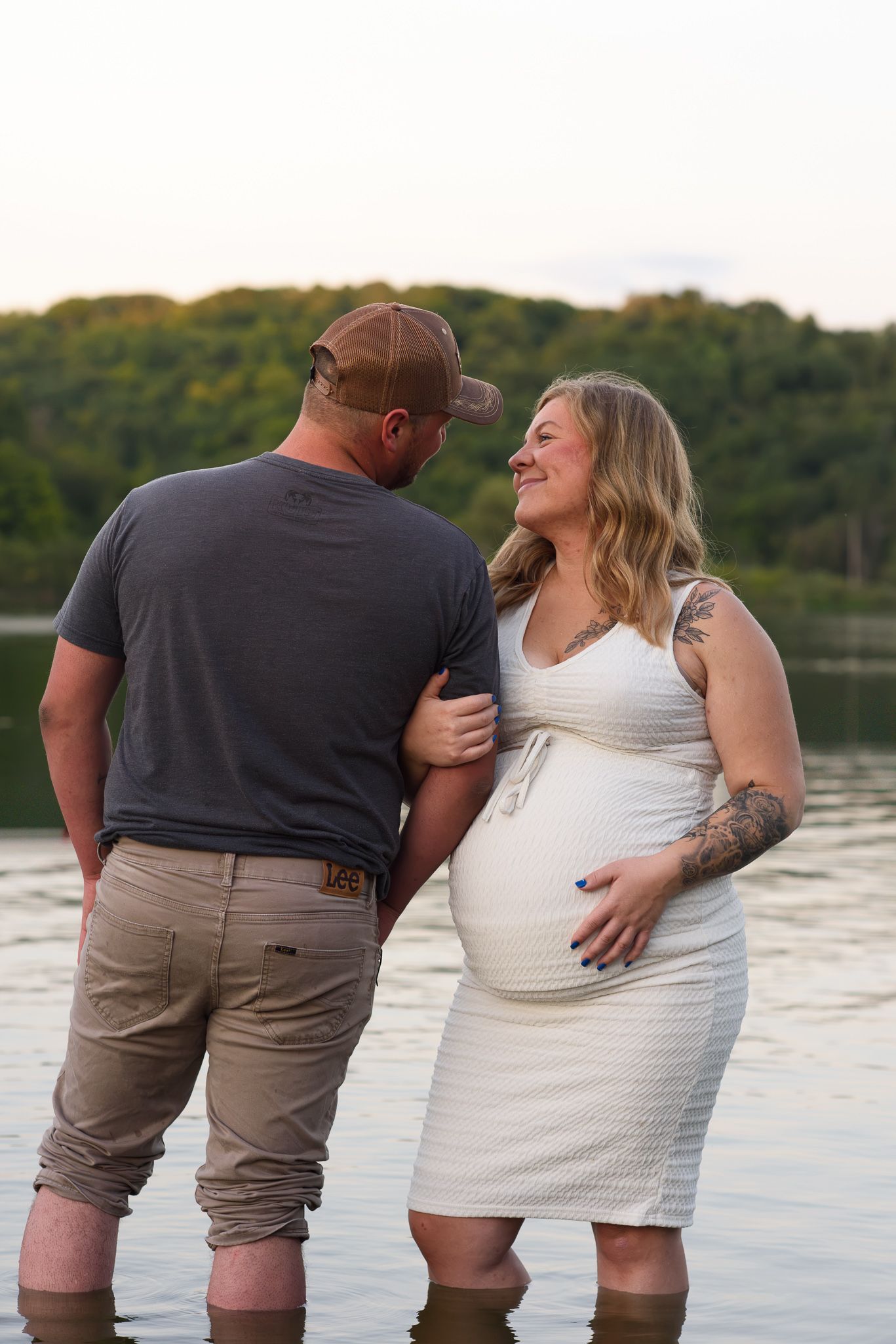 Pregnant couple standing in lake water at sunset, woman in white dress smiling at partner wearing gray shirt and cap.