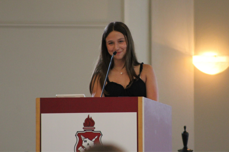 Smiling young woman speaks at a podium during a formal school leadership conference.