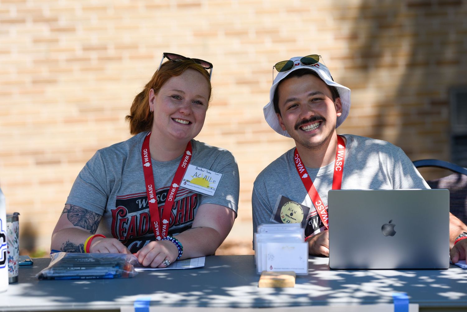 Two smiling volunteers wearing red lanyards and name badges sit at an outdoor registration table with a laptop.