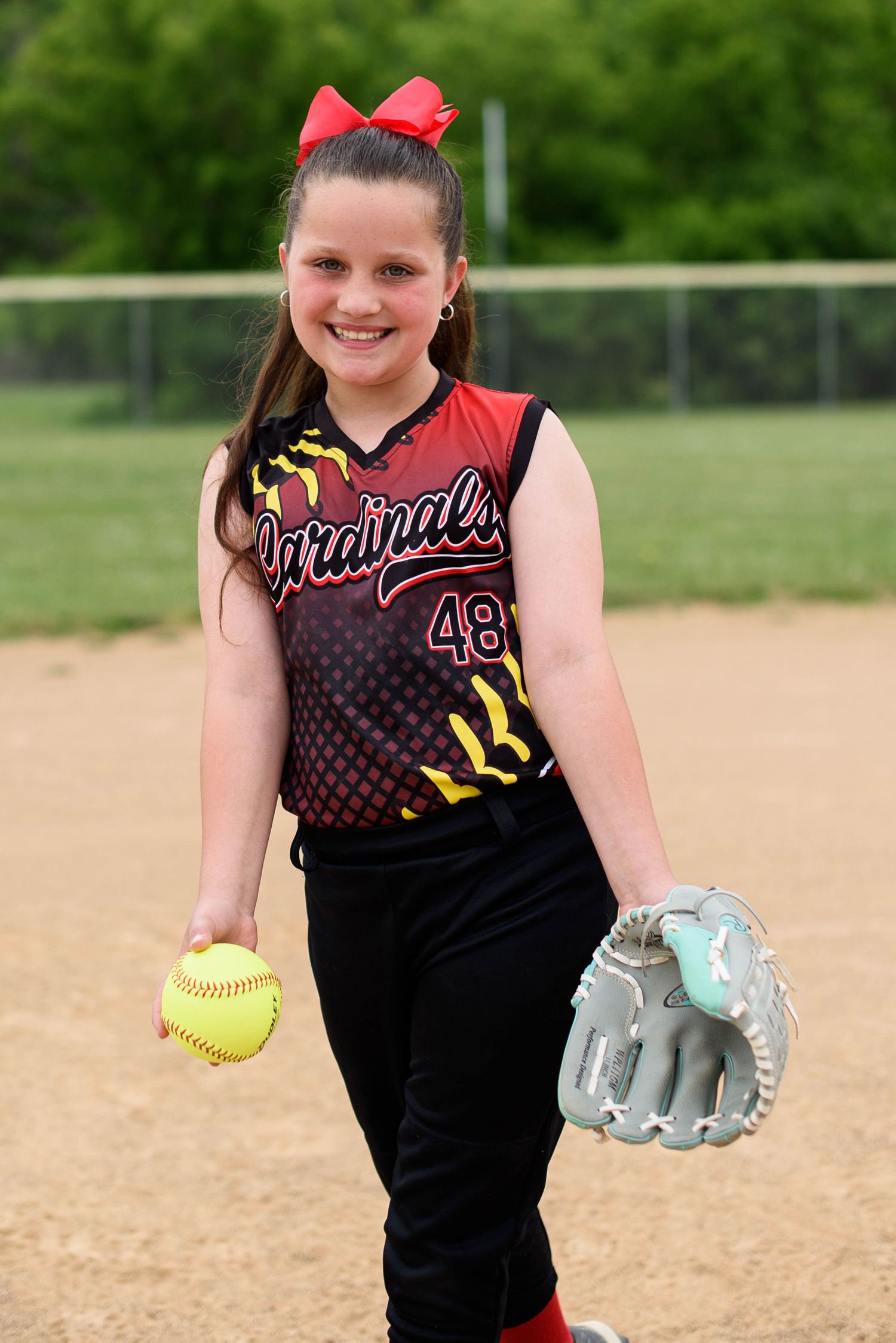 Smiling girl in Cardinals softball uniform holding yellow ball and glove with red bow in hair.