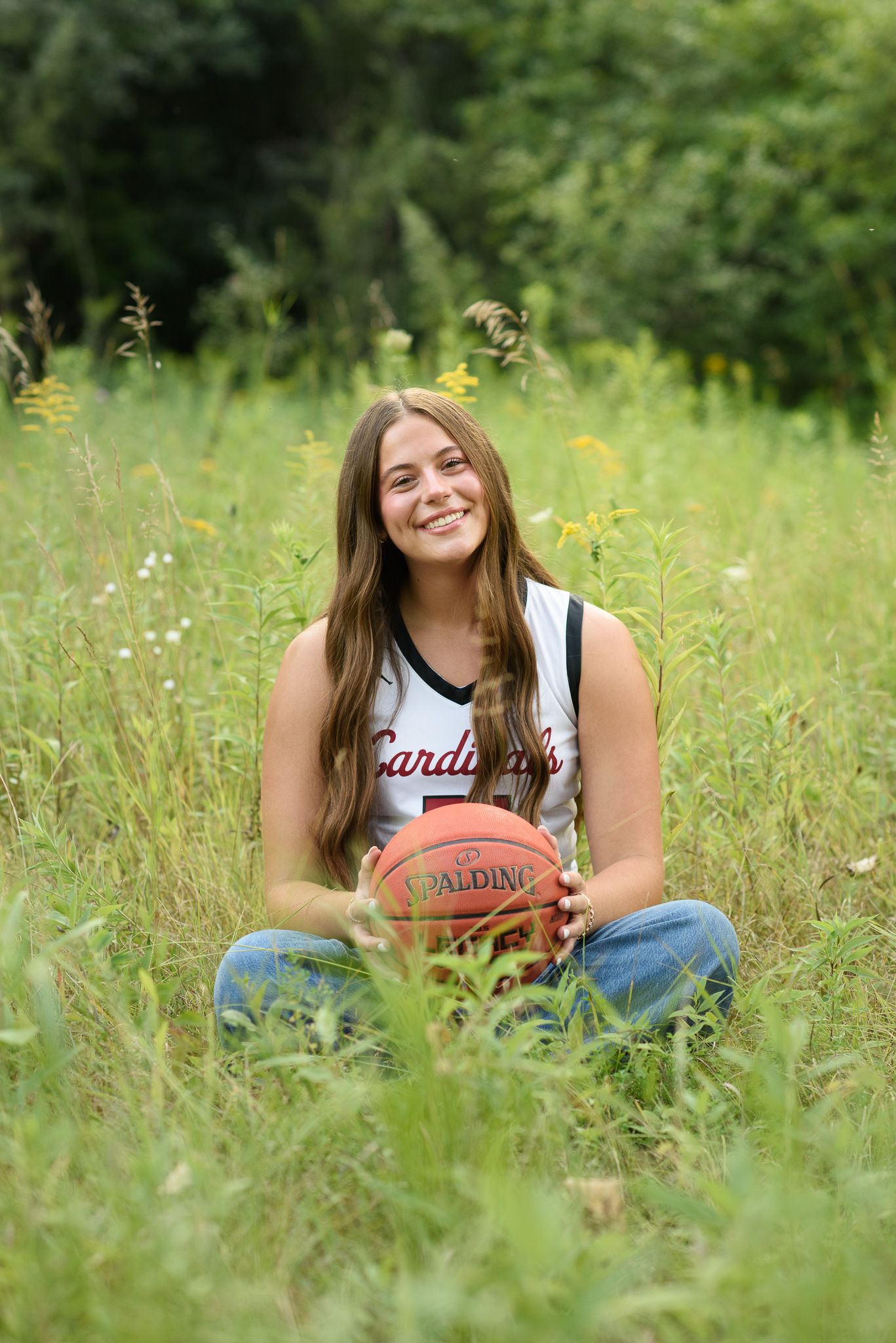 Smiling teenage girl in Cardinals jersey holding Spalding basketball while sitting in a lush green meadow.