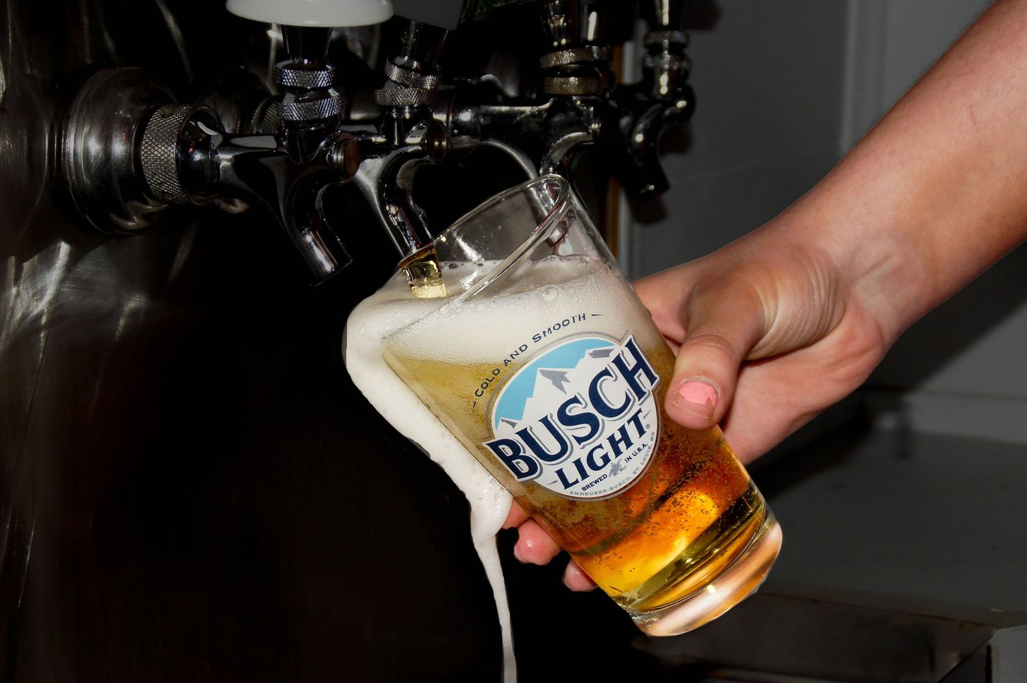 A glass of Busch Light beer overflowing with foam being poured from a tap at a bar.