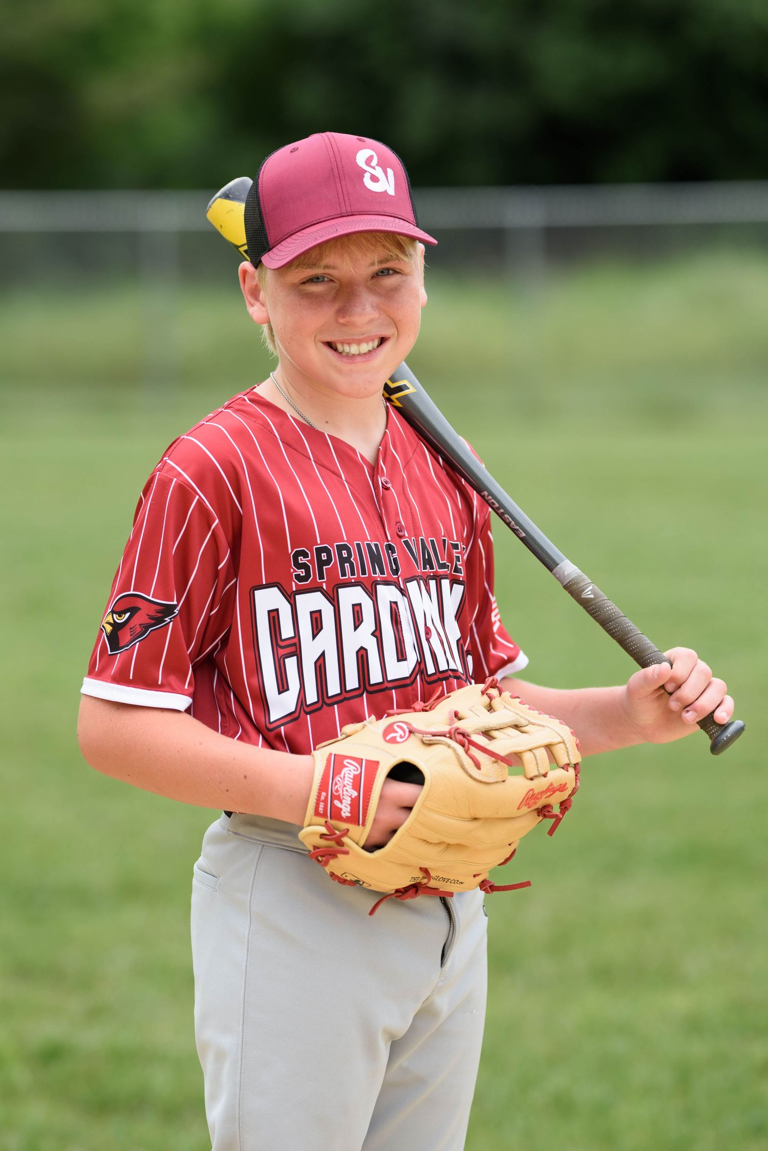 Smiling young Spring Vale Cardinals baseball player poses with bat over shoulder and glove on sunny field.