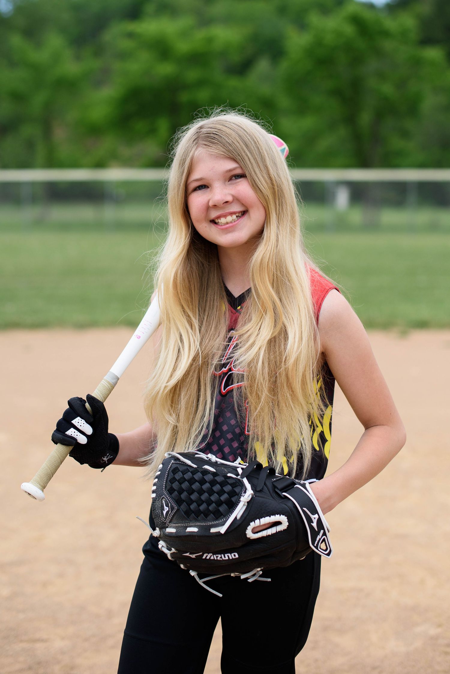 Smiling young softball player holding a bat and glove on a dirt field wearing a black and red jersey.