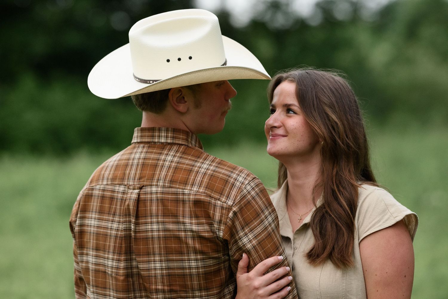 A couple in a green field, man wearing a cowboy hat and plaid shirt, woman smiling warmly at him.