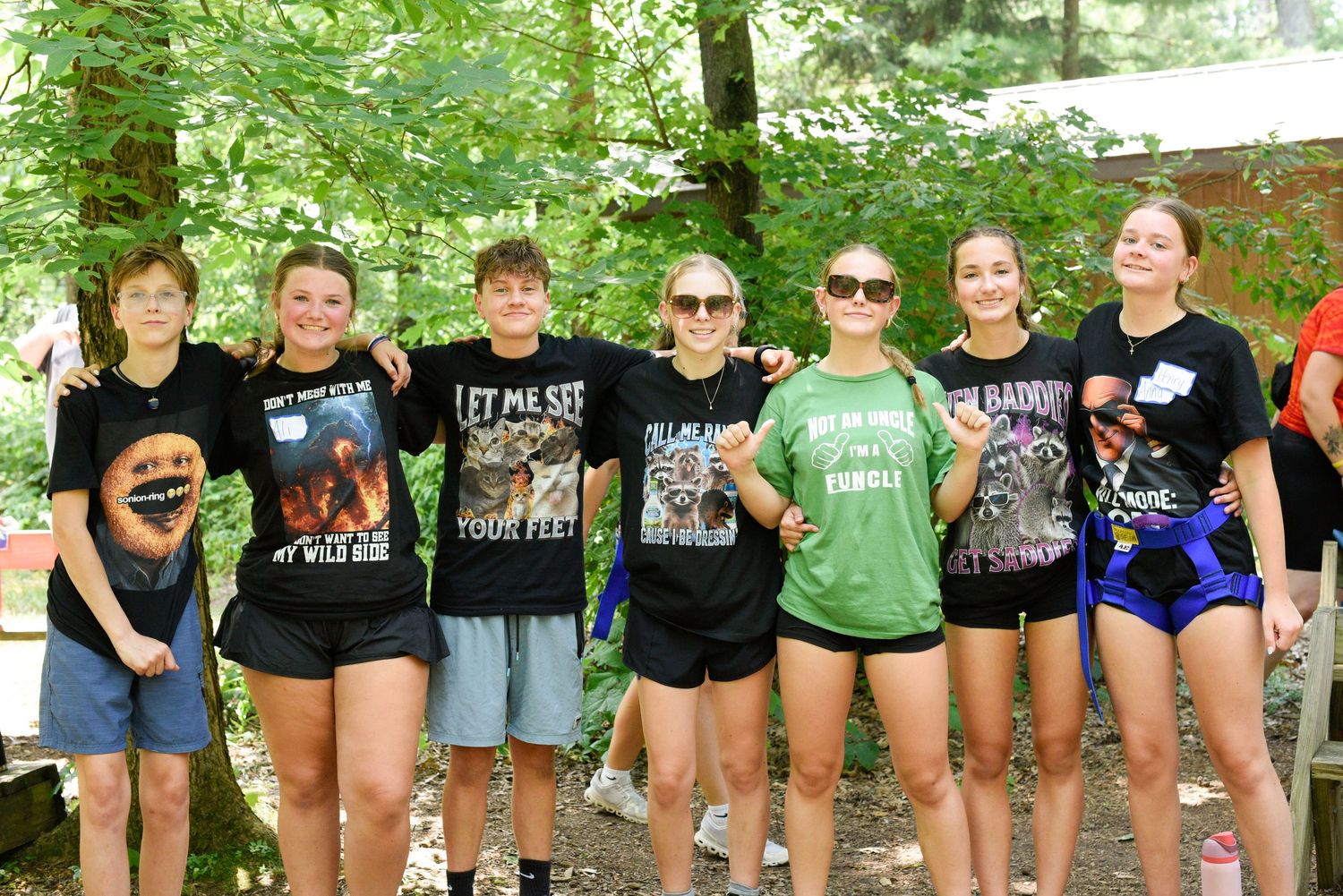 Seven teens pose together smiling in graphic tees at an outdoor summer camp surrounded by trees.