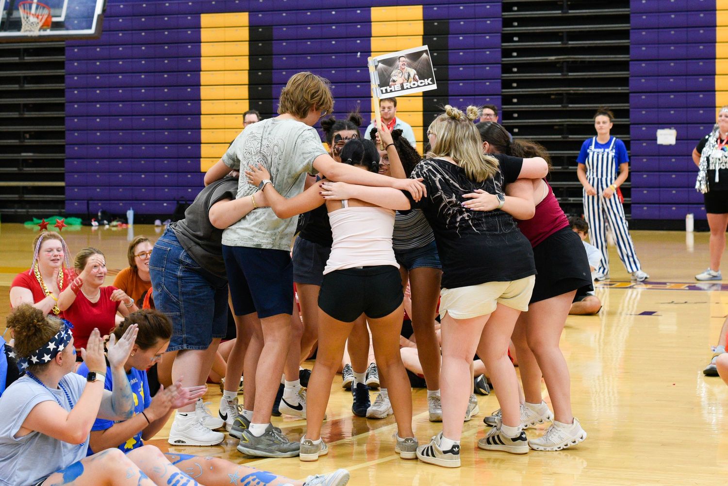 Students celebrate in a group huddle on a gymnasium court during a school event in a purple-accented gym.