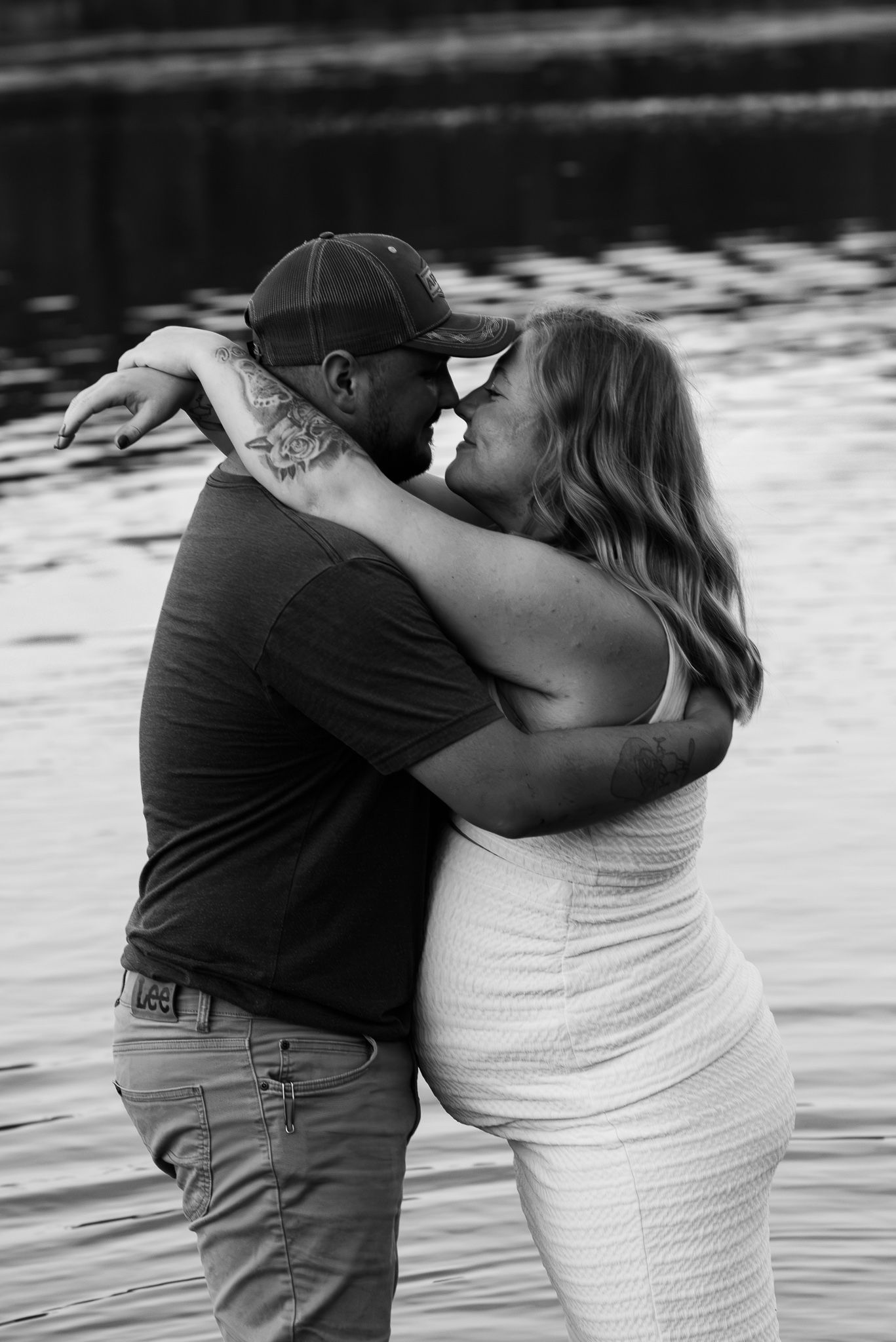 Black and white photo of a couple embracing and smiling near a reflective body of water, man wearing a cap and tattoos.