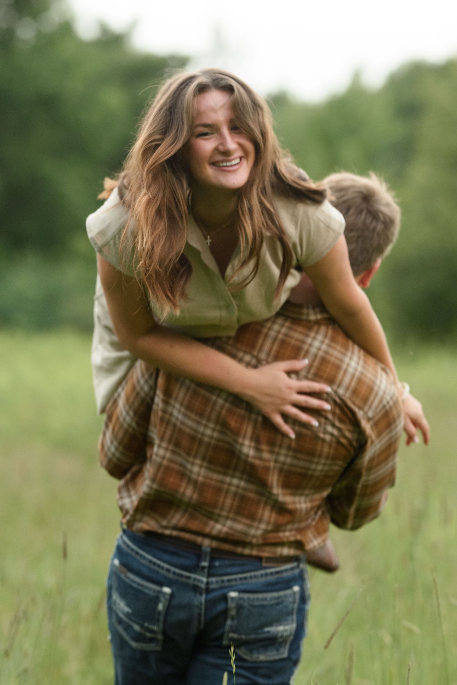 Smiling woman riding piggyback on man in brown plaid shirt through lush green meadow.